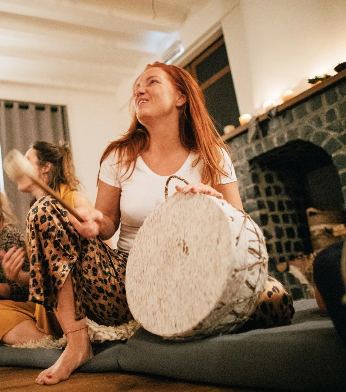 Women sitting on a cushion in a cozy living room, holding a large drum, with a fireplace and candles in the background, enjoying a gathering. music drum instrument muziek singing circle kirtan