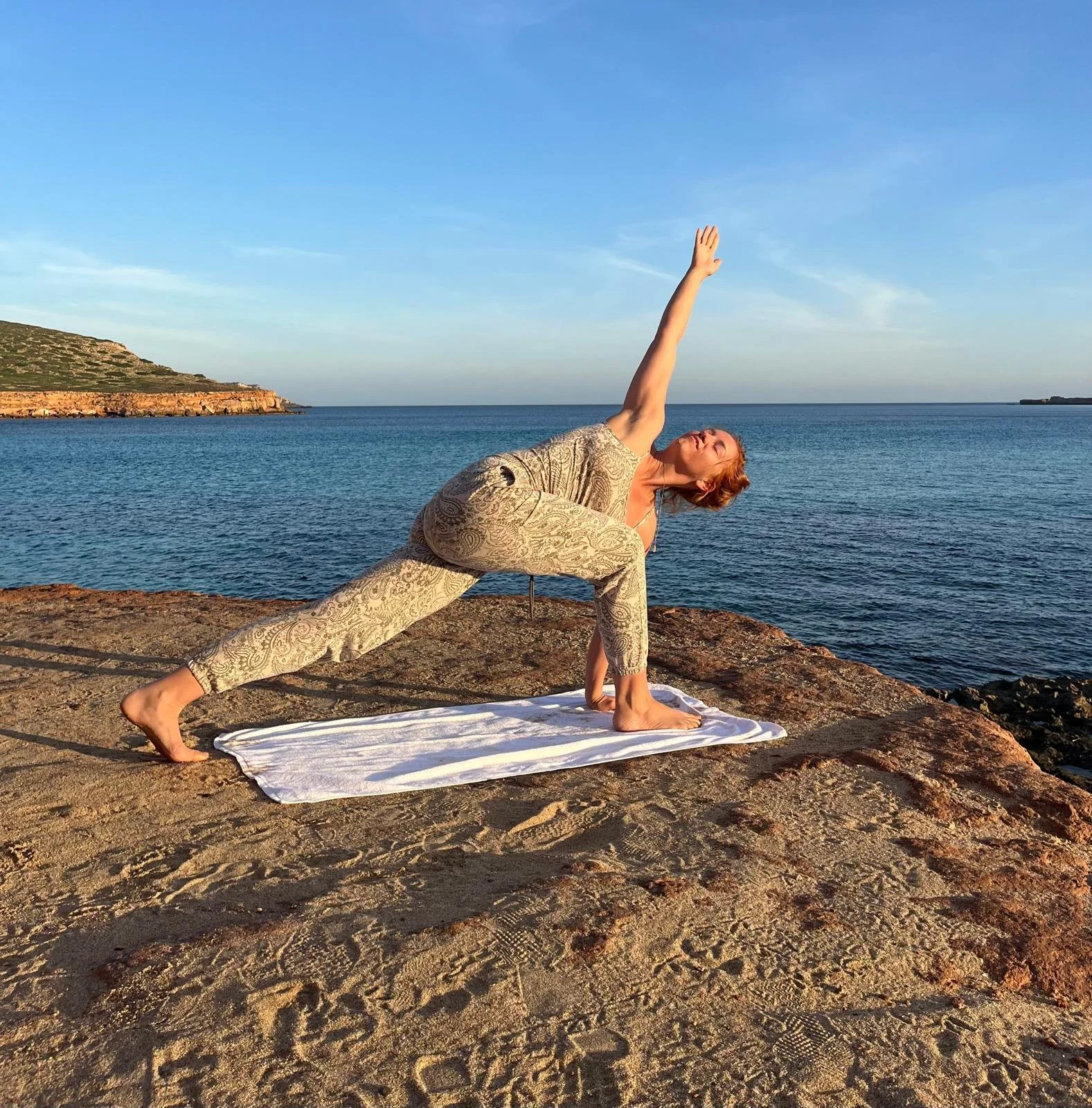 A woman practicing yoga outdoors on a rocky surface near the water, performing a side plank pose with one arm extended upward during sunset or sunrise. lichaamswerk Michelle Kessels teaching facilitator Michelle Ariel tantra teacher embodiment