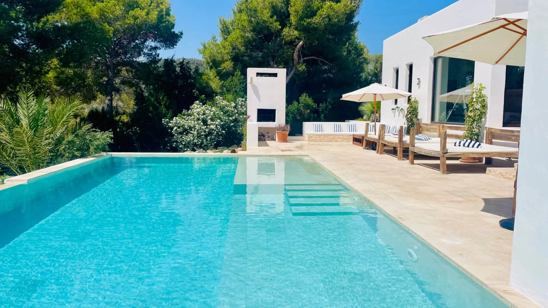 A modern backyard with a turquoise swimming pool, white house, outdoor seating with cushions, potted plants, umbrellas, and lush green trees in the background under a clear blue sky.