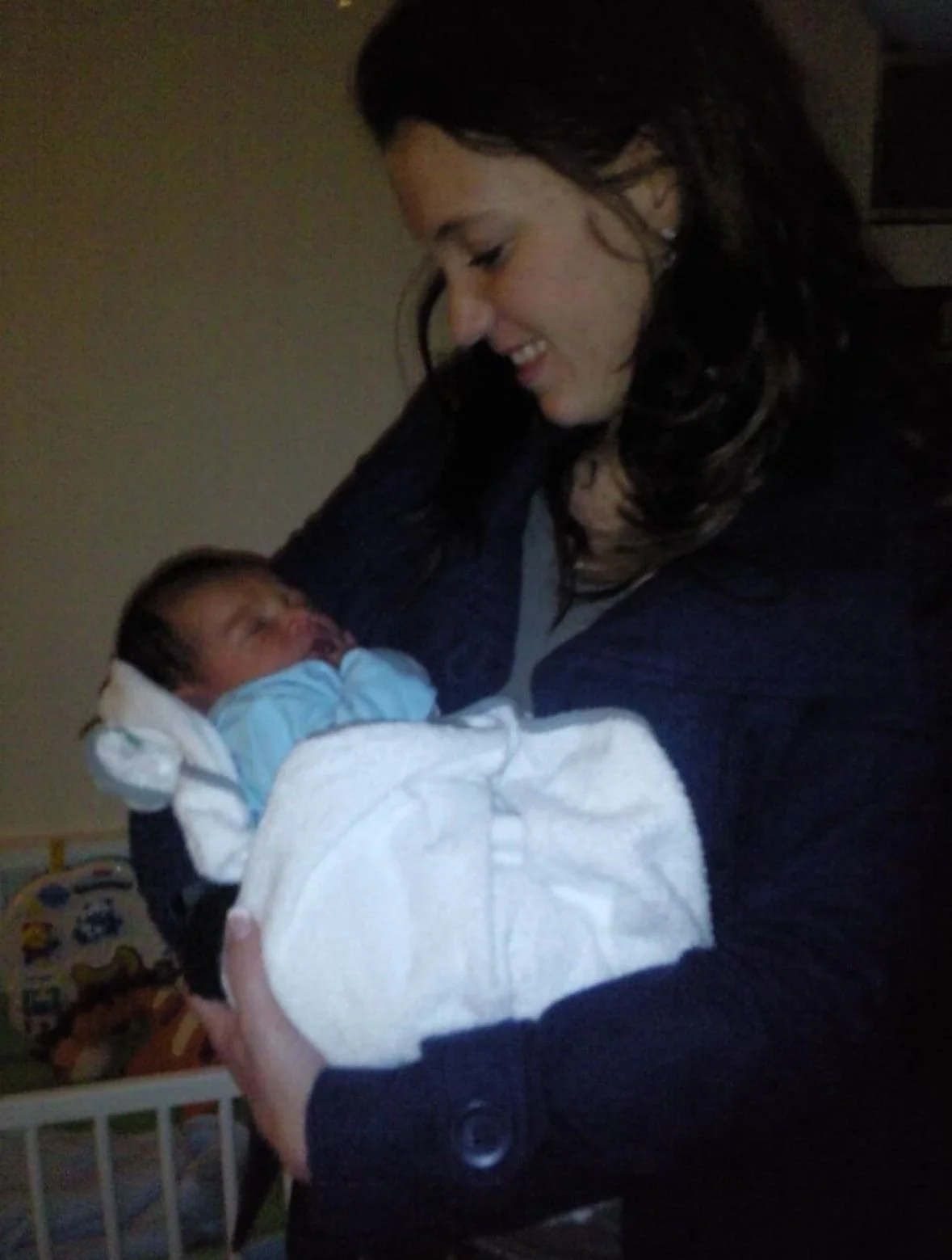 A woman holding a newborn baby in her arms, smiling and looking at the baby, in a dimly lit room. godmother family community