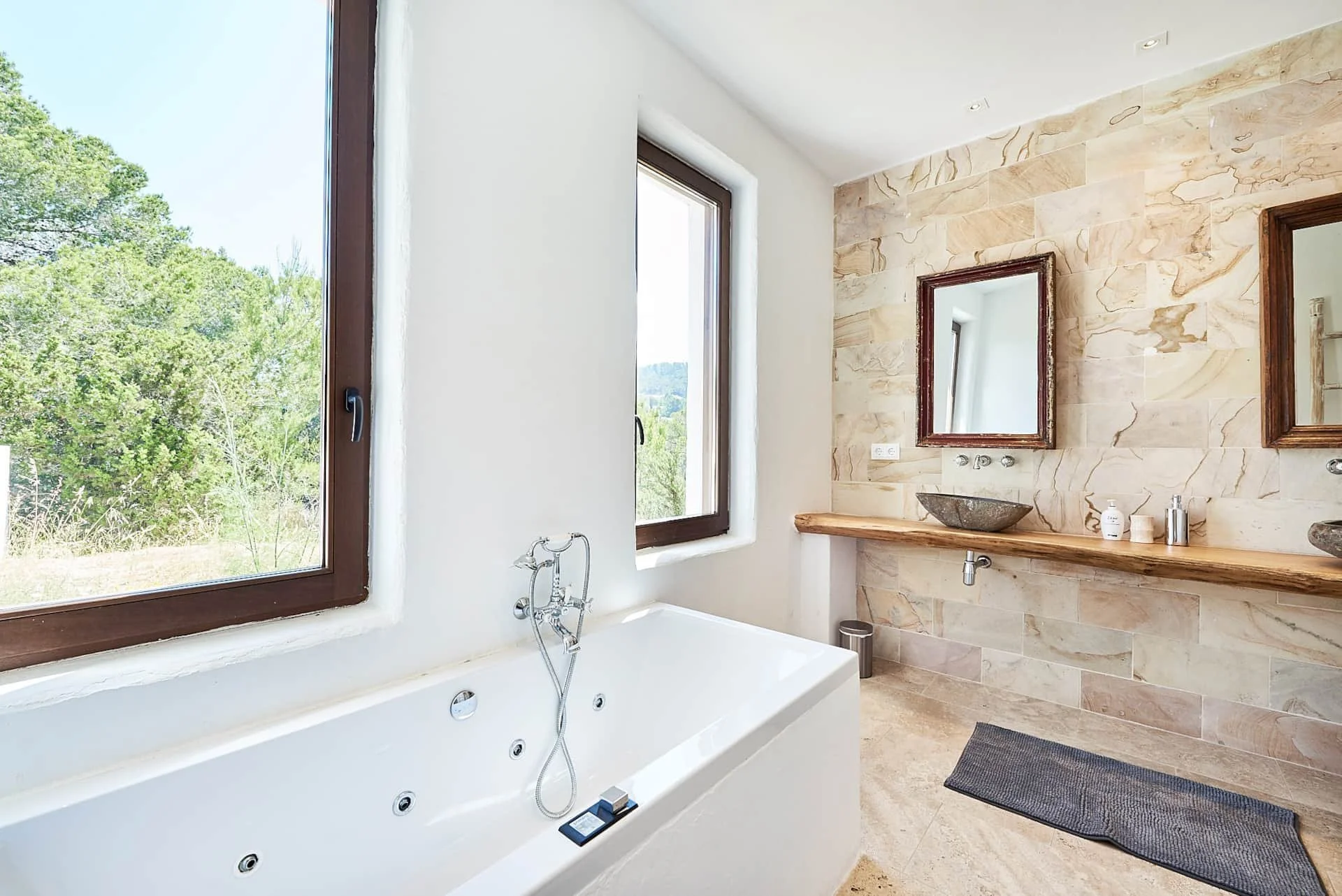Modern bathroom with a large white bathtub, two windows showing greenery outside, a stone wall with two mirrors above a wooden countertop, vessel sinks, and bathroom accessories.