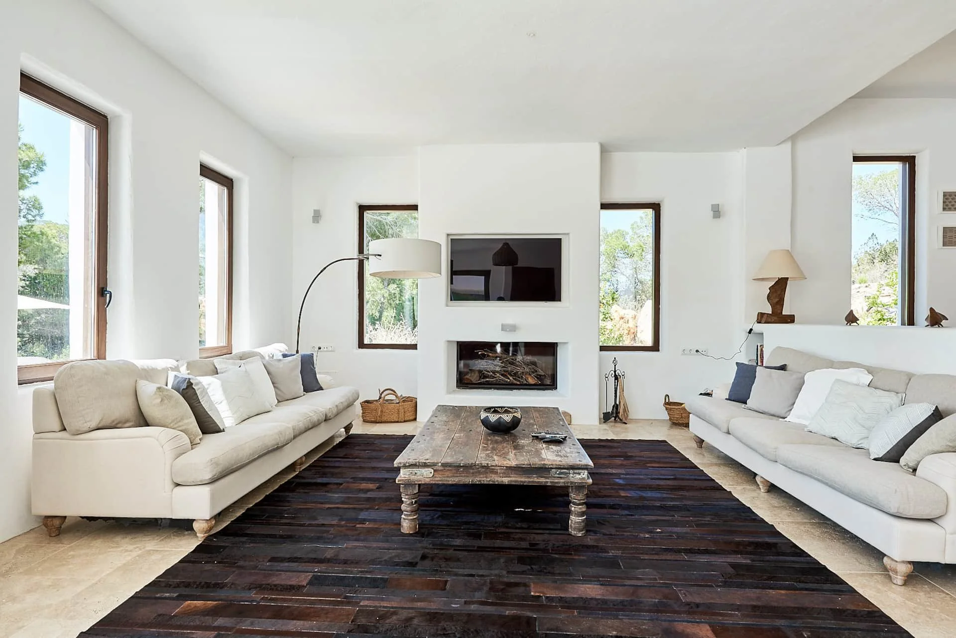 Living room with white walls, large windows, two white sofas with multiple pillows, a rustic wooden coffee table, a black rug, a fireplace with a TV above, decorative lamps, baskets, and greenery outside the windows.
