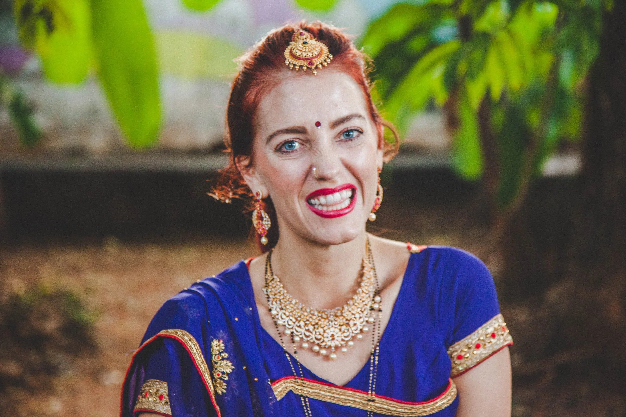 A woman with bright red hair and blue eyes, wearing traditional Indian jewelry and a royal blue dress, smiling outdoors with green foliage in the background. indian traditions