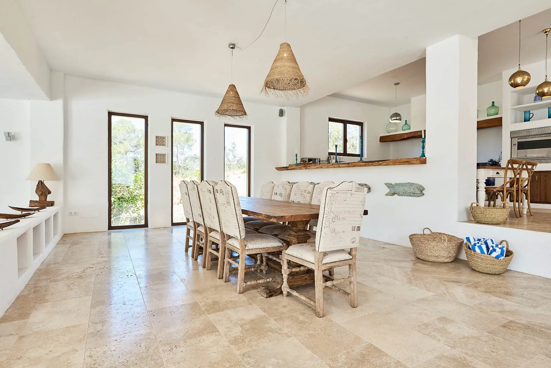 A bright, airy dining room with a large wooden table and eight upholstered chairs, natural light from glass doors and windows, pendant lights hanging from the ceiling, and coastal-themed decorations.