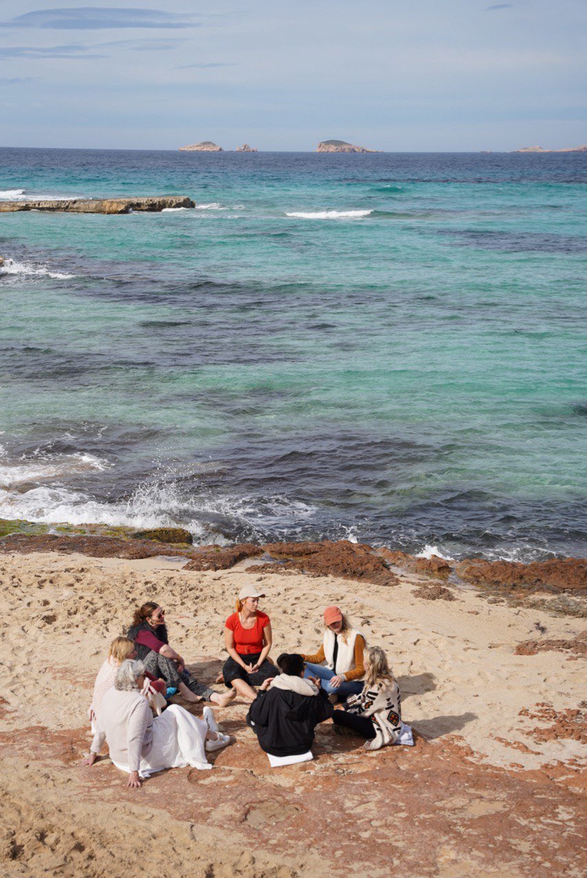 A group of people sitting on a sandy beach near the water, with the ocean and small islands visible in the background.