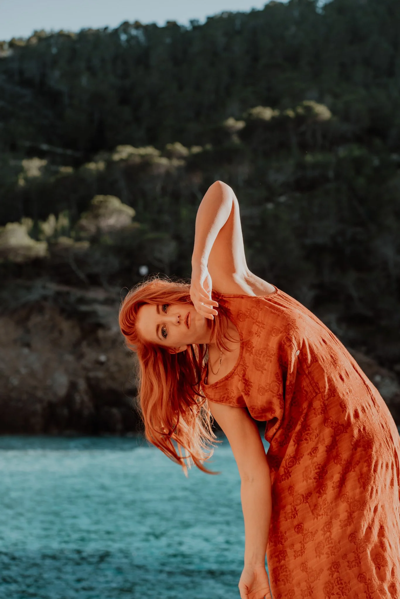 A woman with long red hair wearing an orange dress, standing outdoors near water and a wooded area, leaning to the side with one arm raised above her head. Ibiza beach cala tarida strand rotsen uitzicht turqois sea mountains bergen