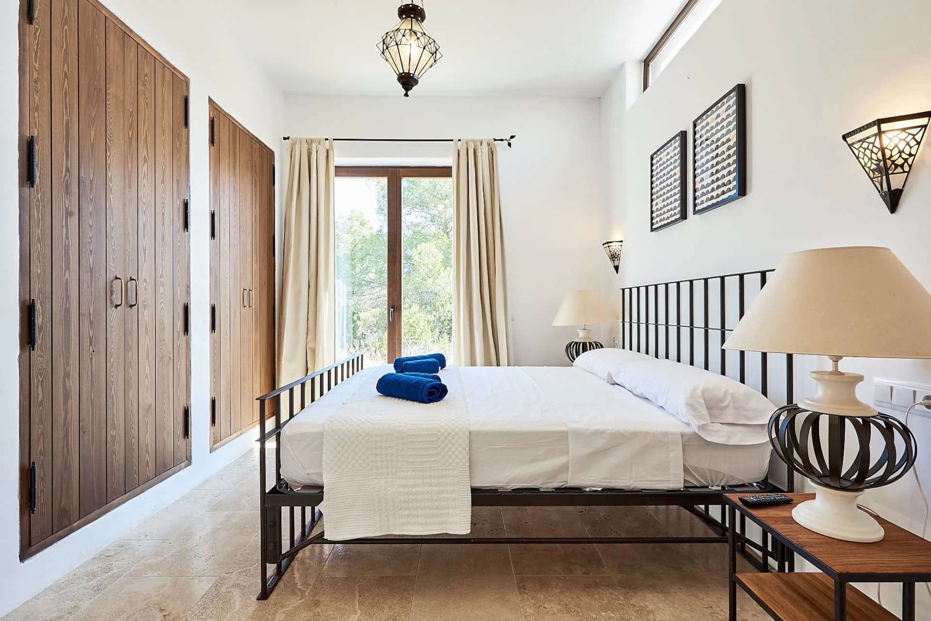 A bedroom with a metal bed frame, white bedding, and blue towels. There is a nightstand with a lamp, two framed pictures on the wall, a large window with curtains, and wooden closet doors.