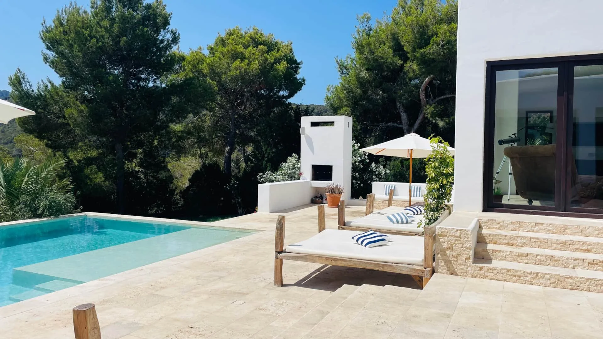 Modern outdoor patio with a swimming pool, lounge chairs with striped pillows, a white umbrella, a white built-in outdoor fireplace, and tall trees in the background on a sunny day.