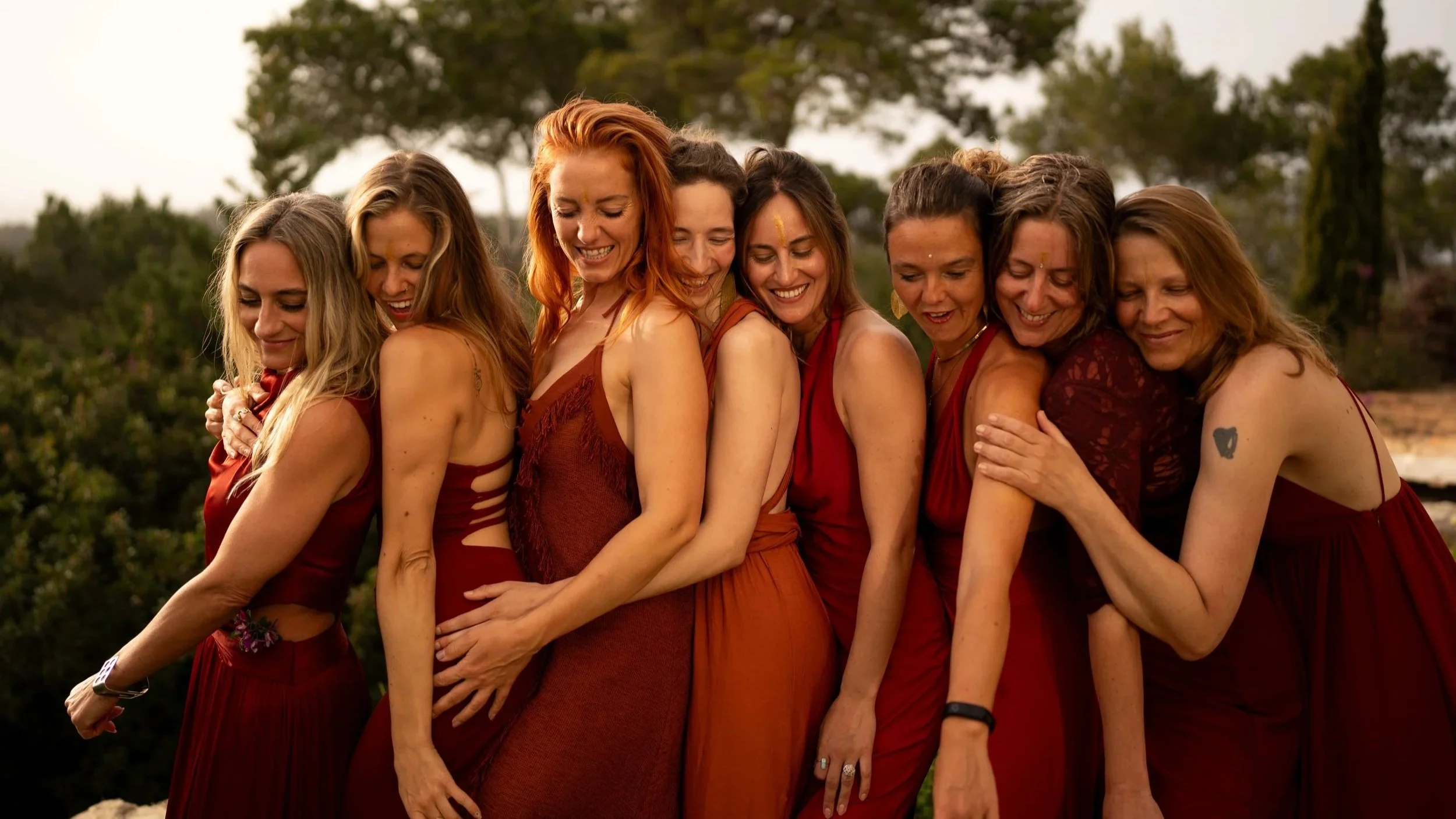 Group of women standing close together outdoors, embracing and smiling, wearing red and orange dresses, with trees and sky in the background.