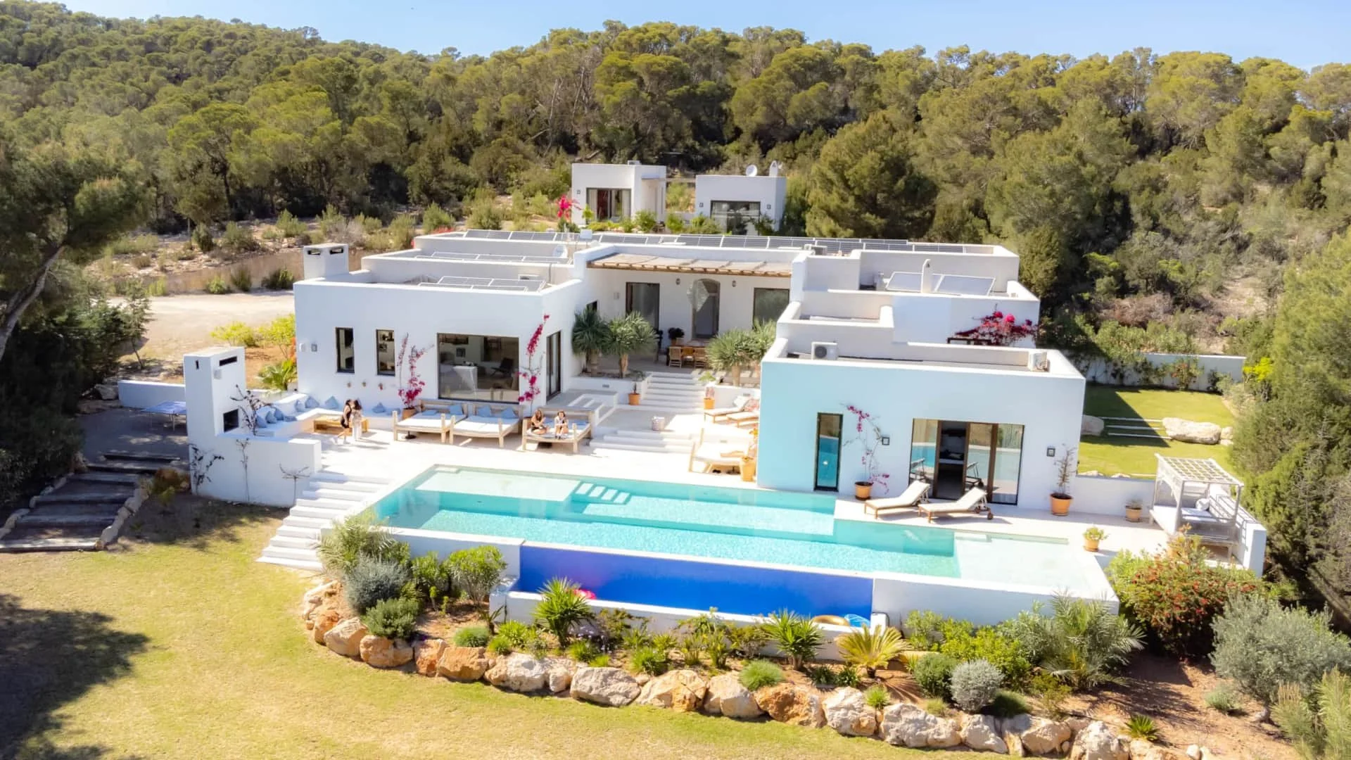 A white modern villa with multiple levels and flat roofs, surrounded by greenery and trees. The villa features an outdoor swimming pool with a stone border, sun loungers, and patio seating areas. There are potted plants and a landscaped garden area around the house.