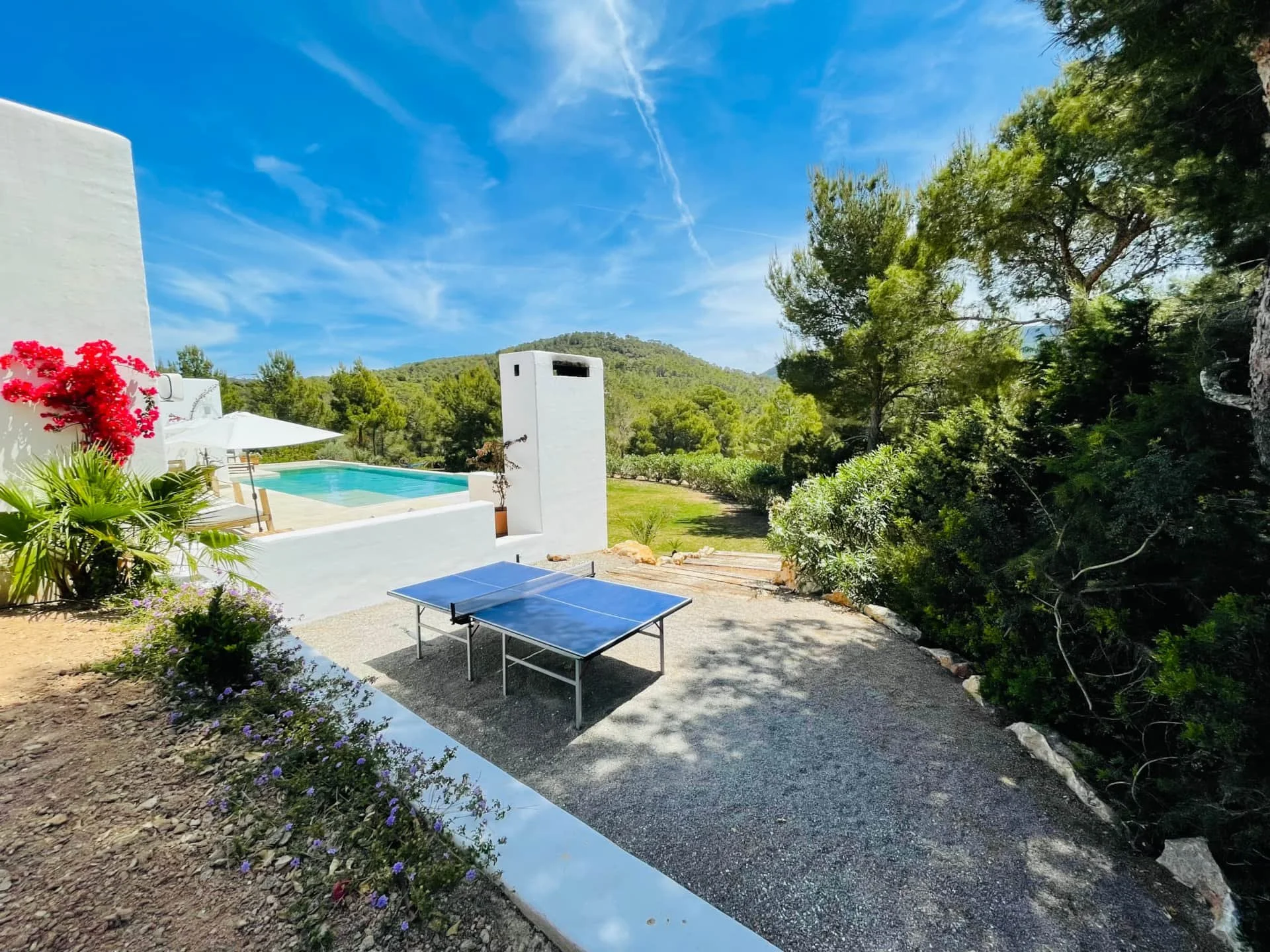 Outdoor patio area with a ping pong table, a swimming pool with white walls, a white umbrella, and lush greenery under a blue sky.