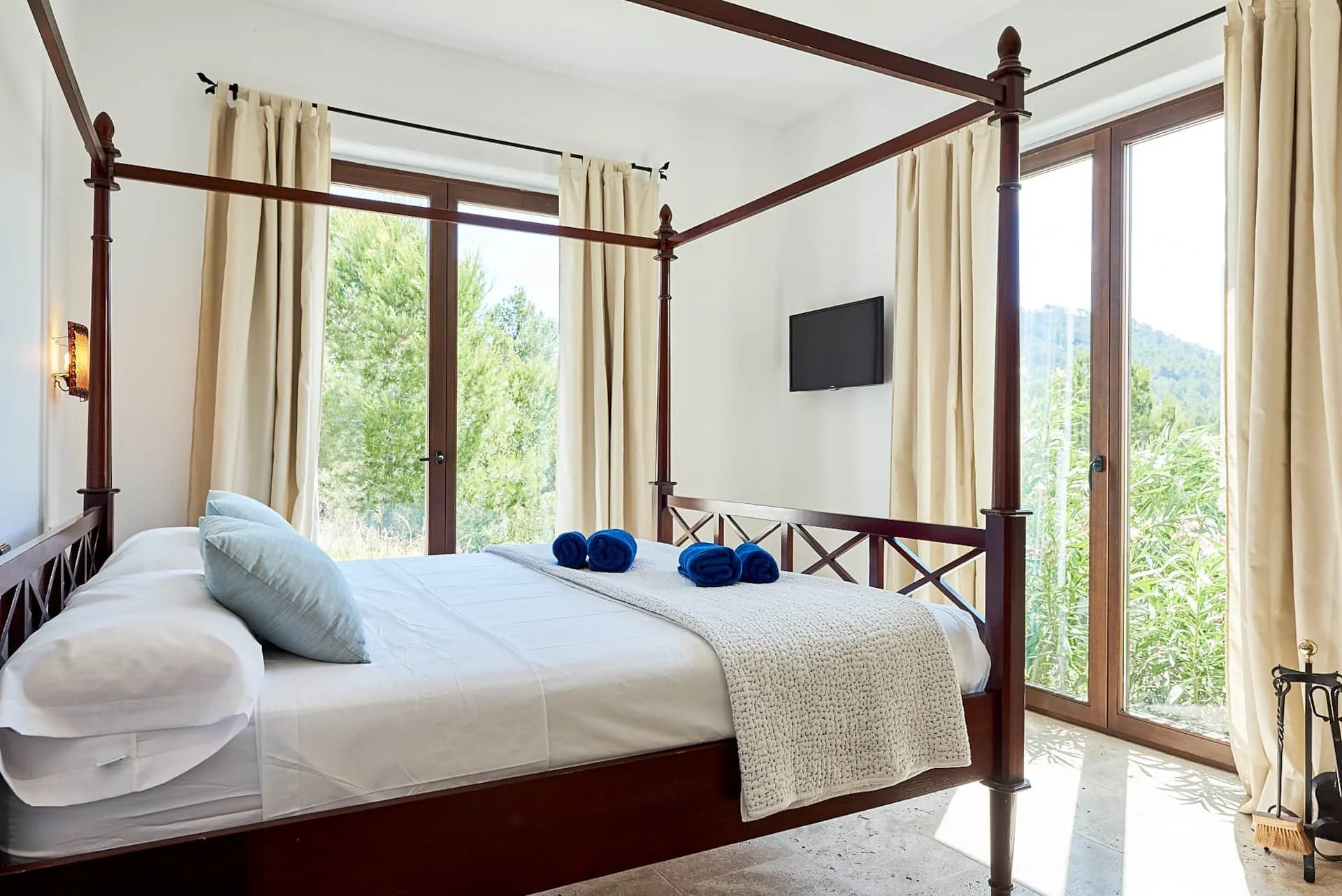 Bedroom with a four-poster bed, white bedding, light blue pillows, and blue rolled towels on top. Large windows with cream curtains, overlooking greenery and hills. A small TV mounted on the wall.
