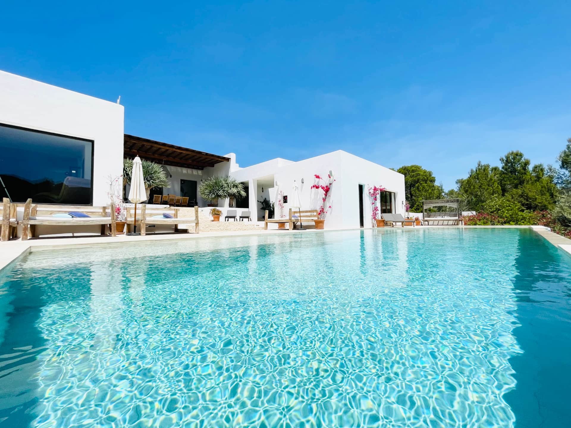 Clear swimming pool in front of white modern villa with outdoor seating, potted plants, and a blue sky.