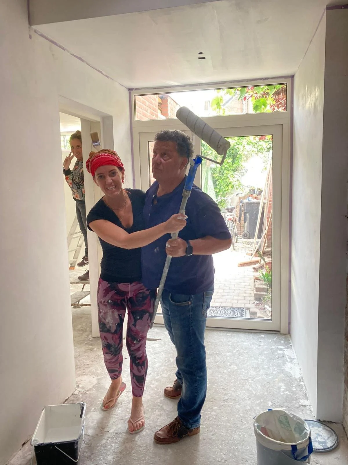 Three people in a room under construction; a woman with a red bandana and patterned pants is smiling, a man with a roller brush is standing with a woman, and another woman is peeking from background. doe het zelver klussen