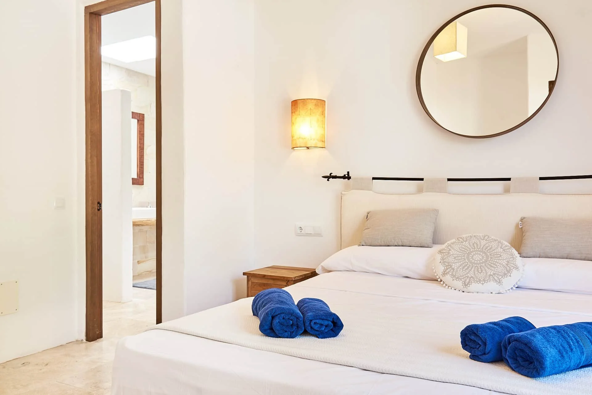 A neatly made bed in a bright, minimalistic bedroom with four blue rolled towels on top, a decorative pillow, two grey pillows, a wooden nightstand, a wall-mounted lamp, a round mirror, and a doorway leading to a bathroom.