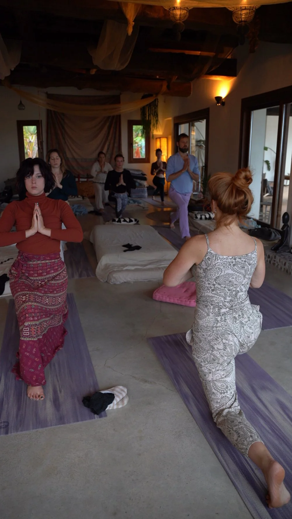 People participating in a yoga or meditation class, some in a kneeling position, inside a cozy, softly-lit room with large windows and wooden ceiling beams.