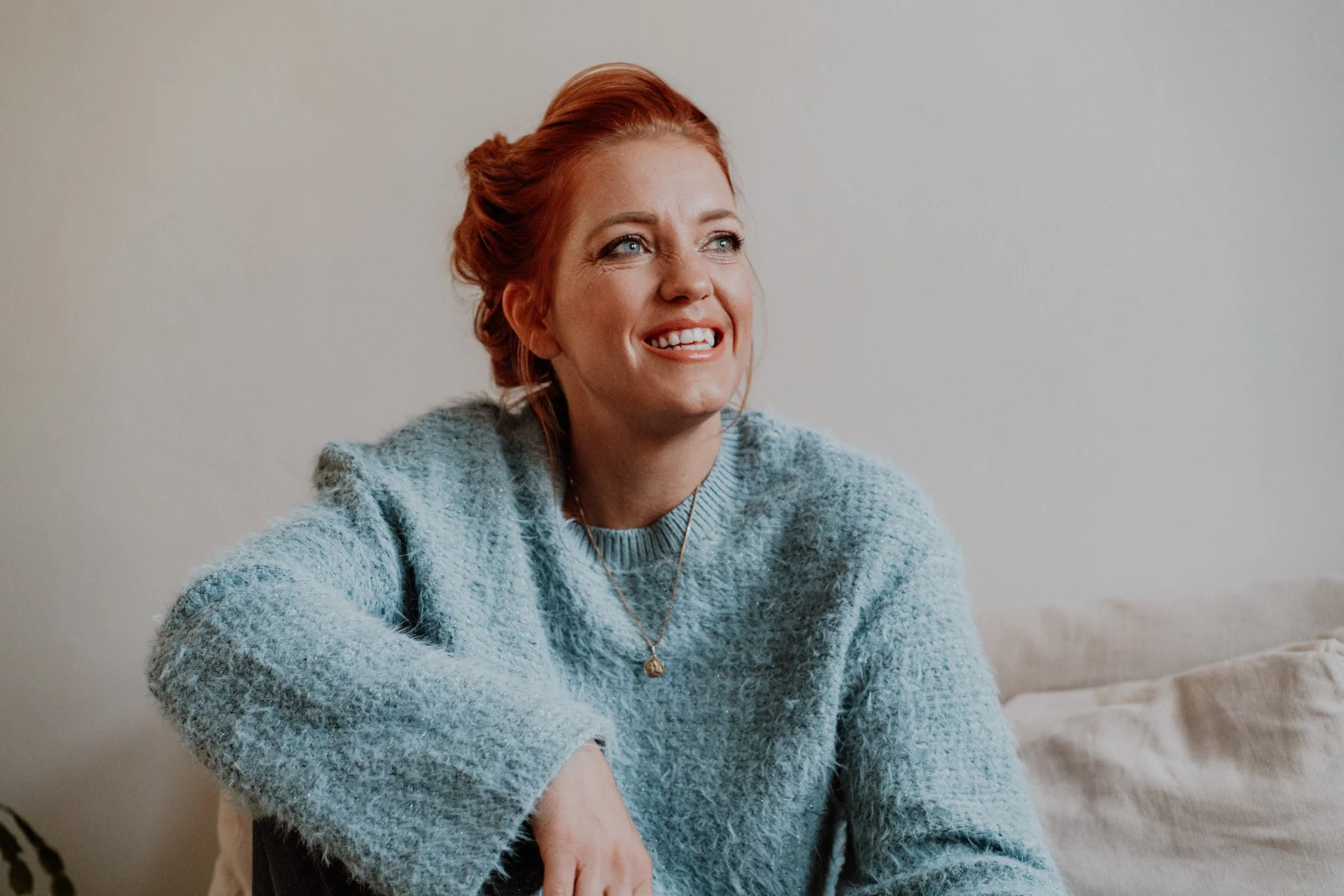 A woman with red hair and blue eyes sitting on a beige couch, wearing a blue fuzzy sweater and smiling while looking to her right.