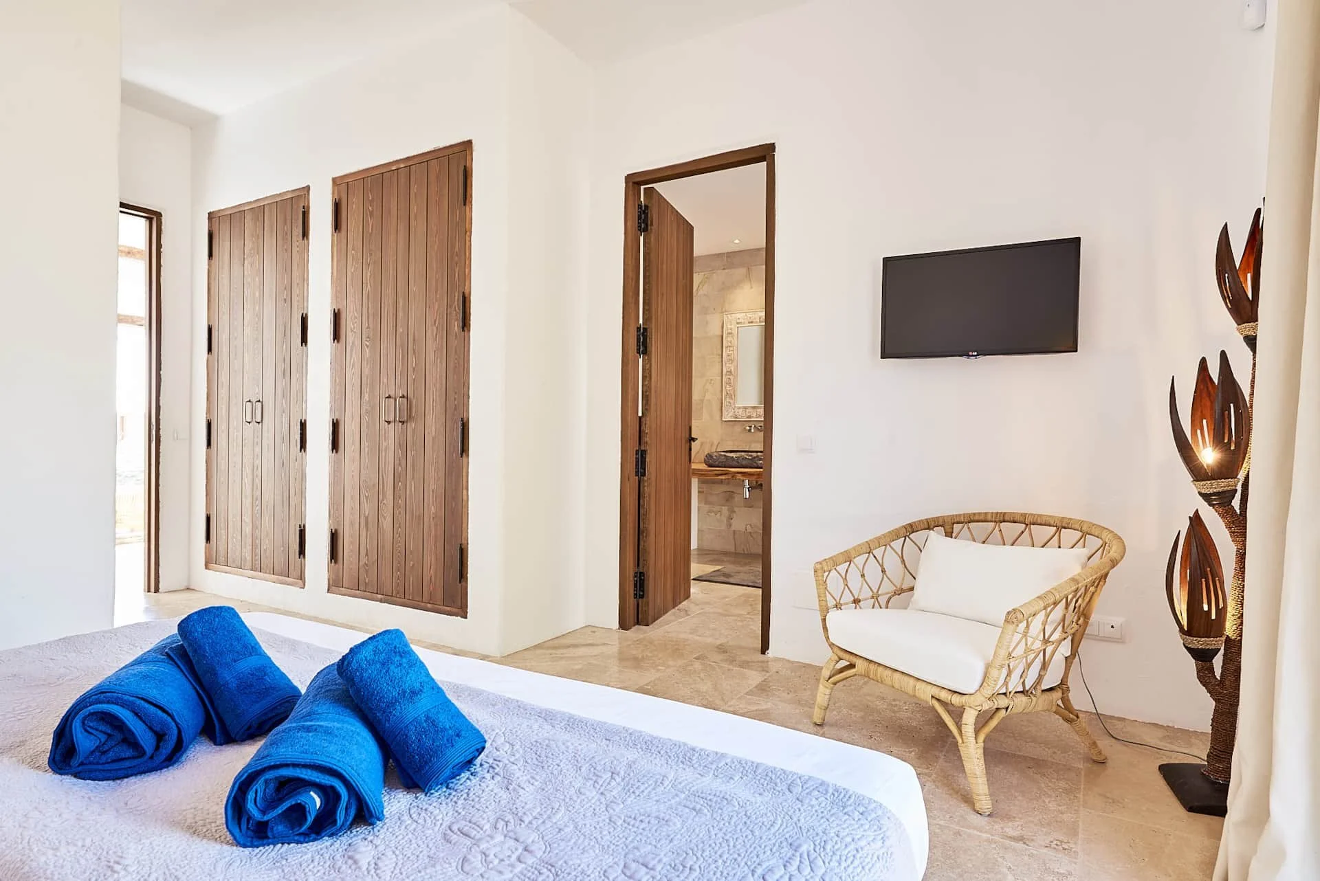 A bedroom with a bed covered with a white quilt and three blue rolled towels on top. There are wooden closet doors, a TV mounted on a white wall, a rattan chair with white cushion, and a tall decorative lamp with leaf-shaped shades. An open door reveals a bathroom with a sink and mirror.