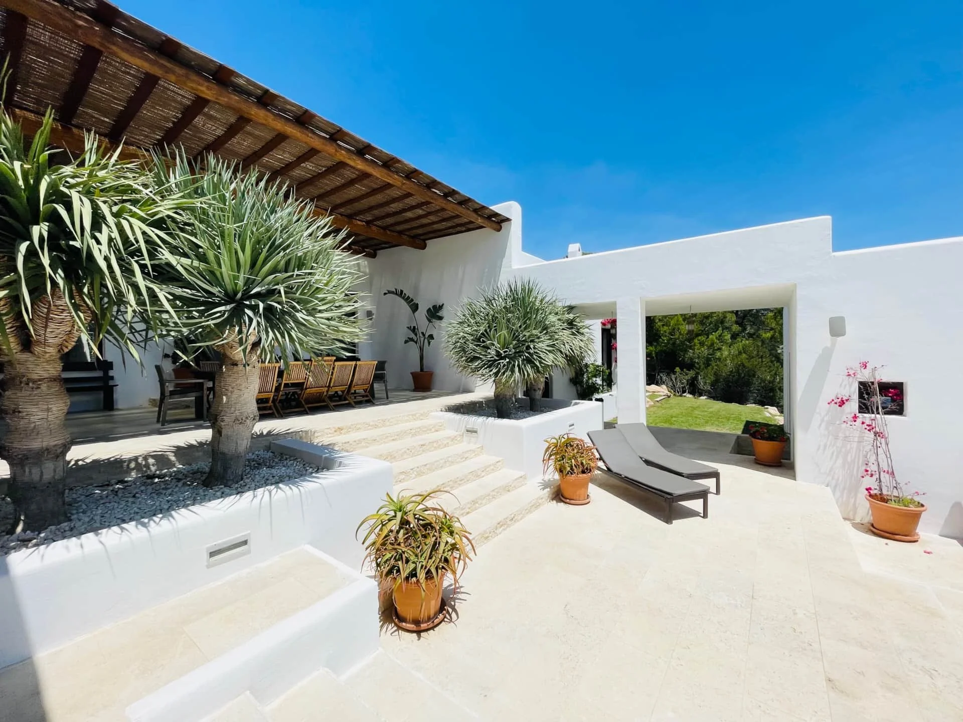 A bright outdoor terrace with white walls, potted plants, and lounge chairs under a wooden pergola, with a clear blue sky and green garden in the background.