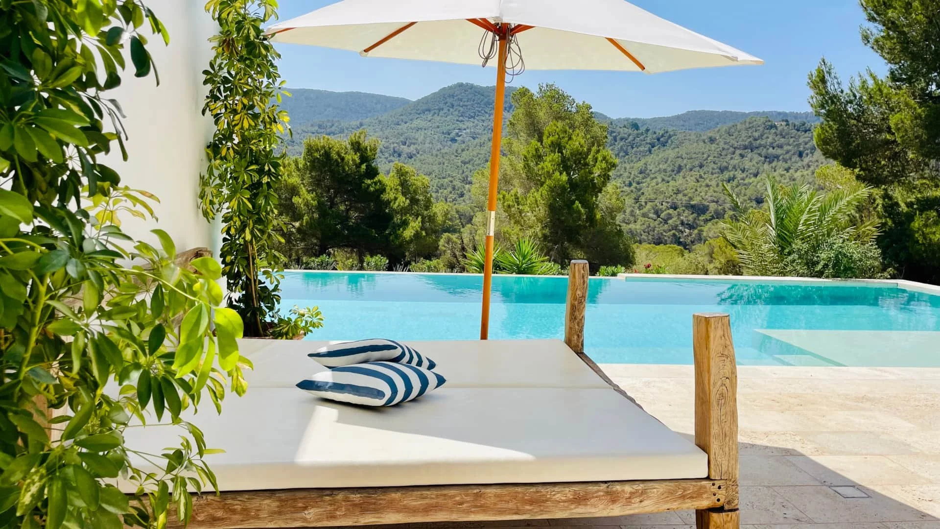 Poolside area with lounge bed and pillows under a large white patio umbrella, surrounded by green trees and mountains in the background.