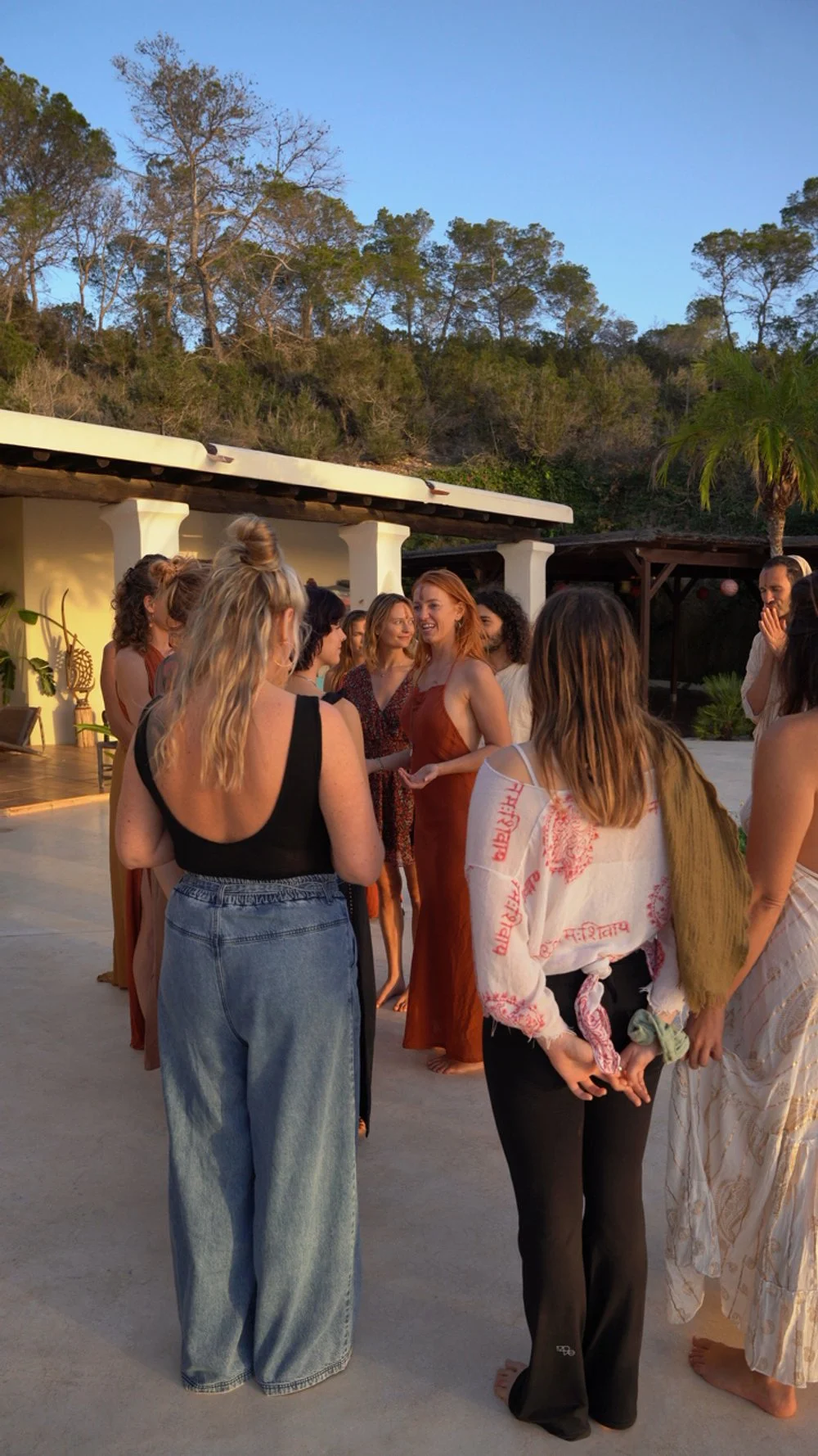 A group of people gathered outdoors, socializing and enjoying a gathering during sunset, with trees and a building with white columns in the background.