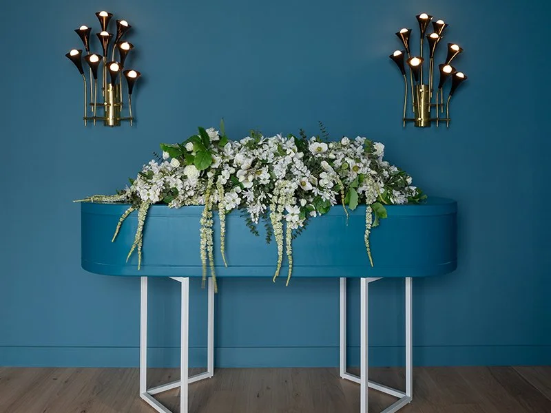 A modern blue bespoke casket decorated with a large white floral arrangement.