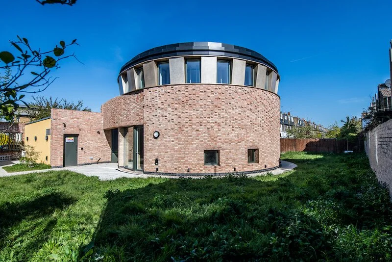 Modern building with a cylindrical brick base and a rounded glass and concrete top, surrounded by a grassy yard.