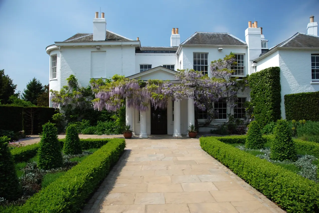 White multi-story house with large windows, surrounded by a green garden with trimmed hedges and flowering trees, under a blue sky.
