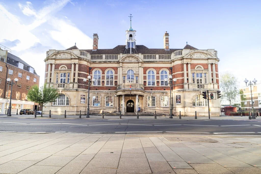 A historic brick and stone building with large arched windows, a central entrance, and a small tower with a weather vane on top, located on a city street with lampposts and traffic lights.