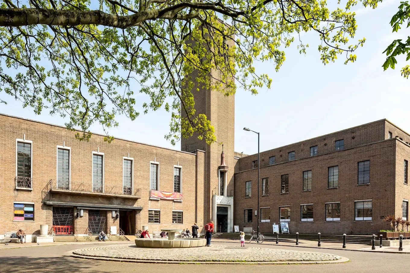 A brick building with a bell tower in a courtyard, with people walking and sitting, and trees with green leaves overhead.