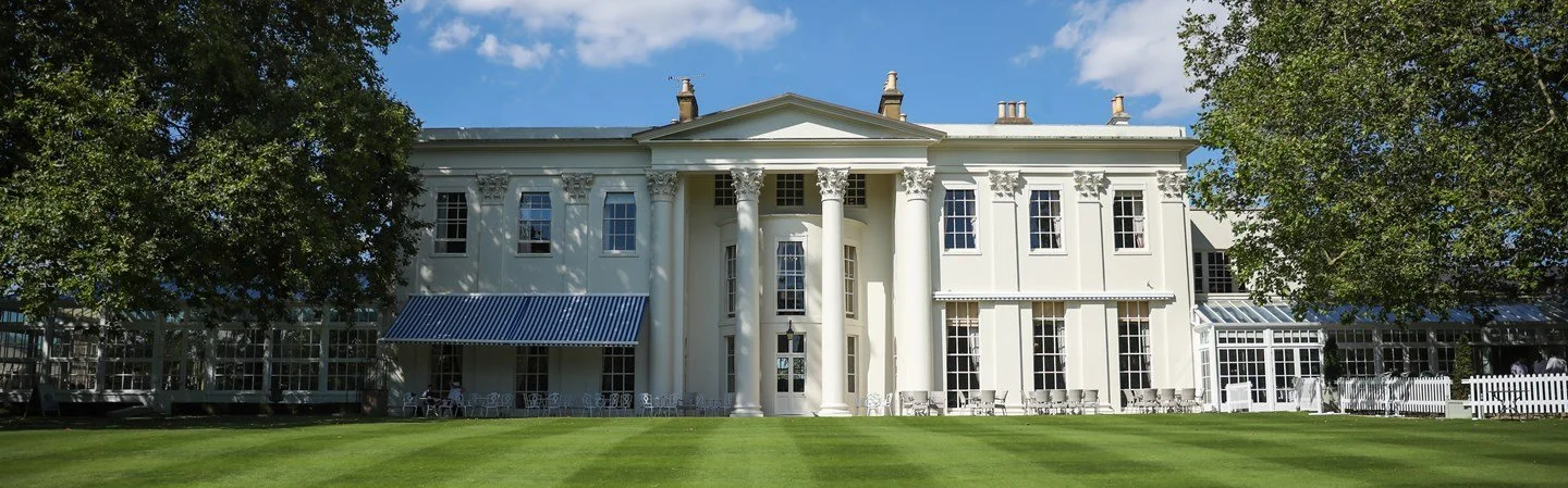 A large white mansion with columns, windows, and a blue striped awning, surrounded by green trees and grass under a clear blue sky.