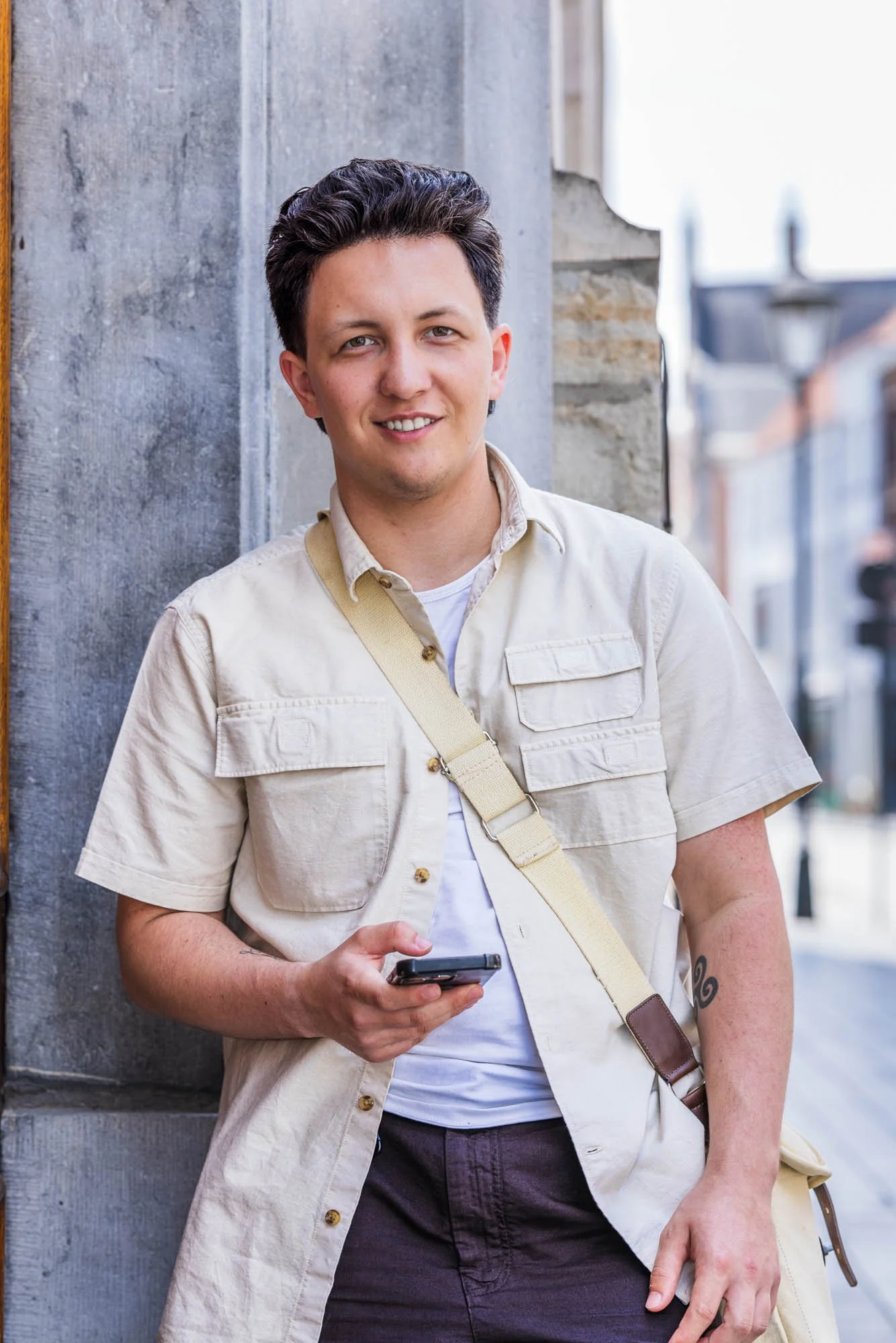 Informele portretfoto van Alex Verspecht leunend tegen een gevel en glimlachend met smartphone in de hand, Mechelen"
