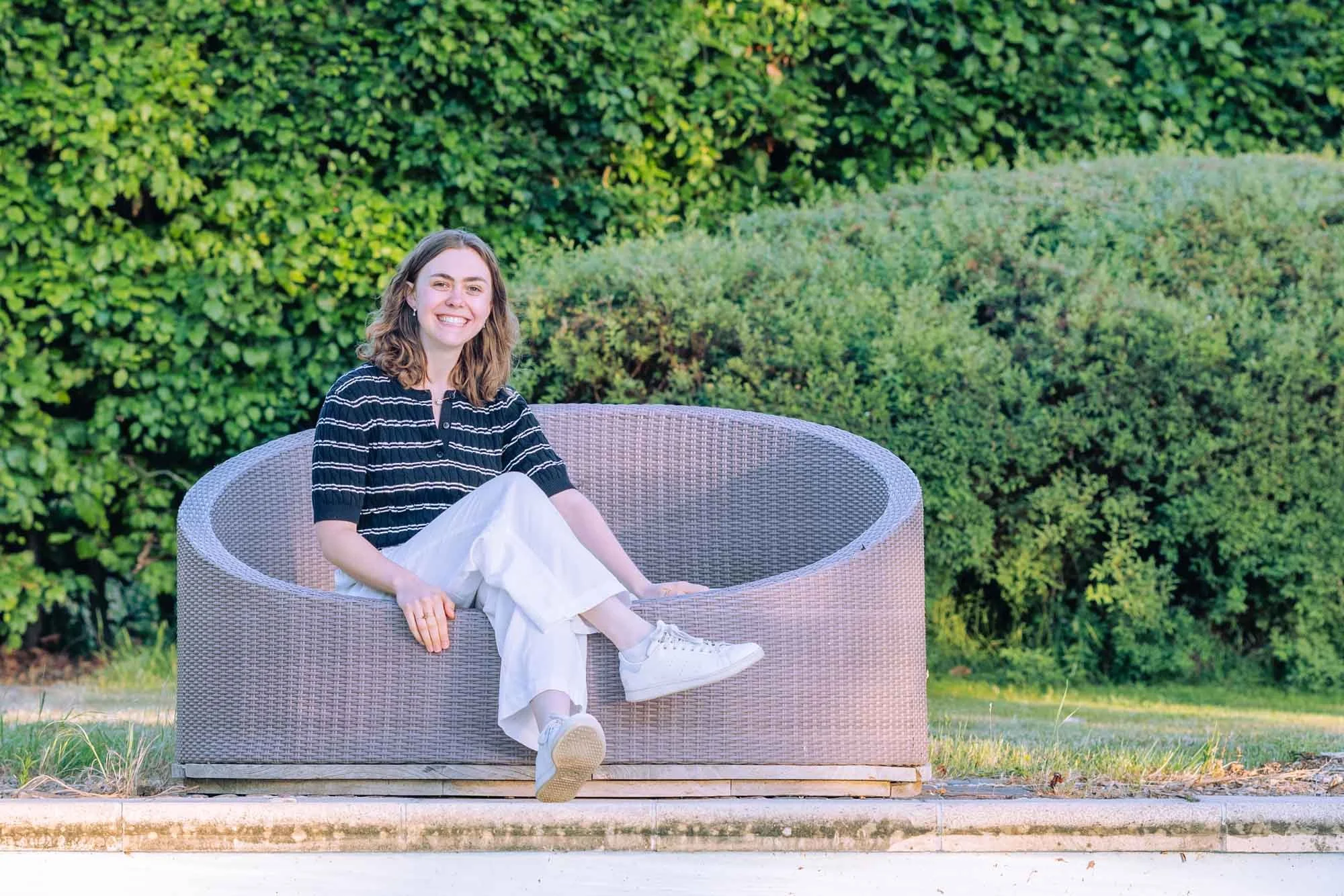 "Soloportret van Amber Sels zittend in een buitenzetel in de tuin, met een grote glimlach en casual houding, gefotografeerd in Rijmenam aan het zwembad"
