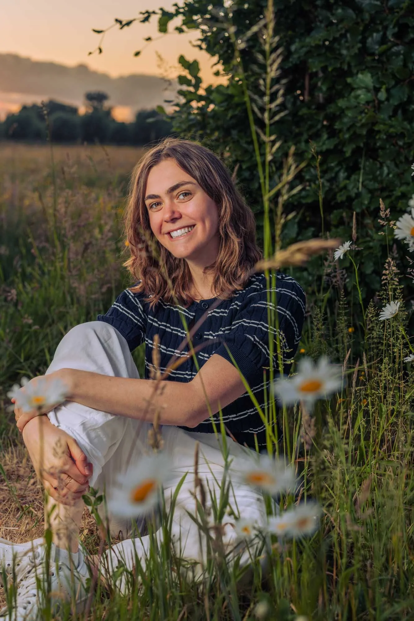 Vrouw zittend in een bloemenveld, kijkend naar de camera bij gouden uur
Jonge vrouw zittend tussen margrieten in een veld bij zonsondergang. Portret door Leysen Photography, met natuurlijke glimlach en warme avondzon.
