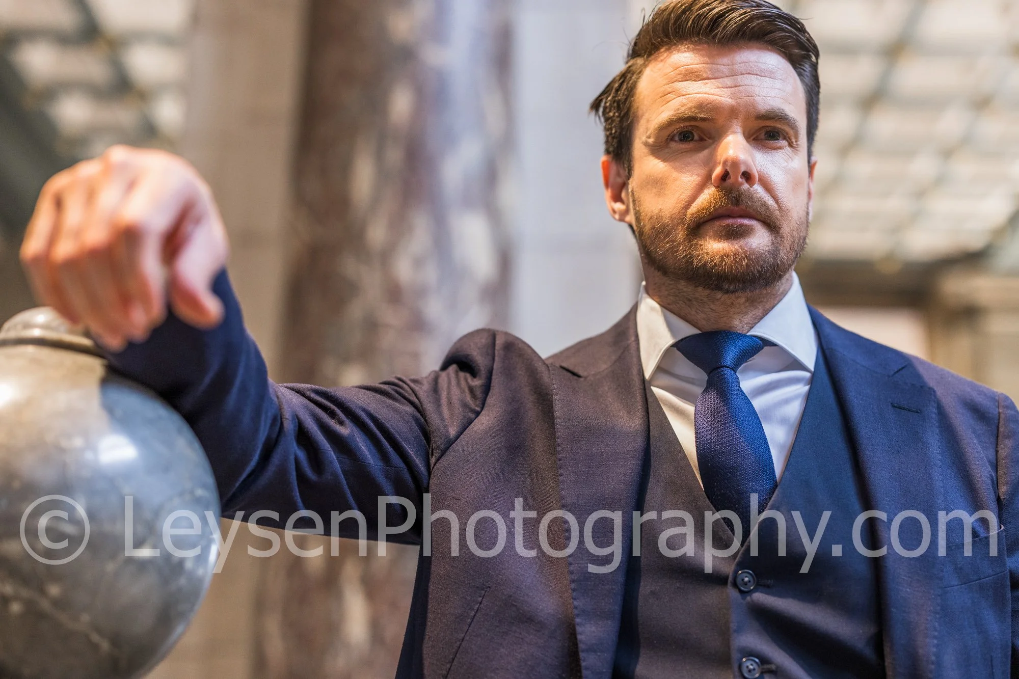 Confident businessman in navy suit leaning on marble railing inside historic building