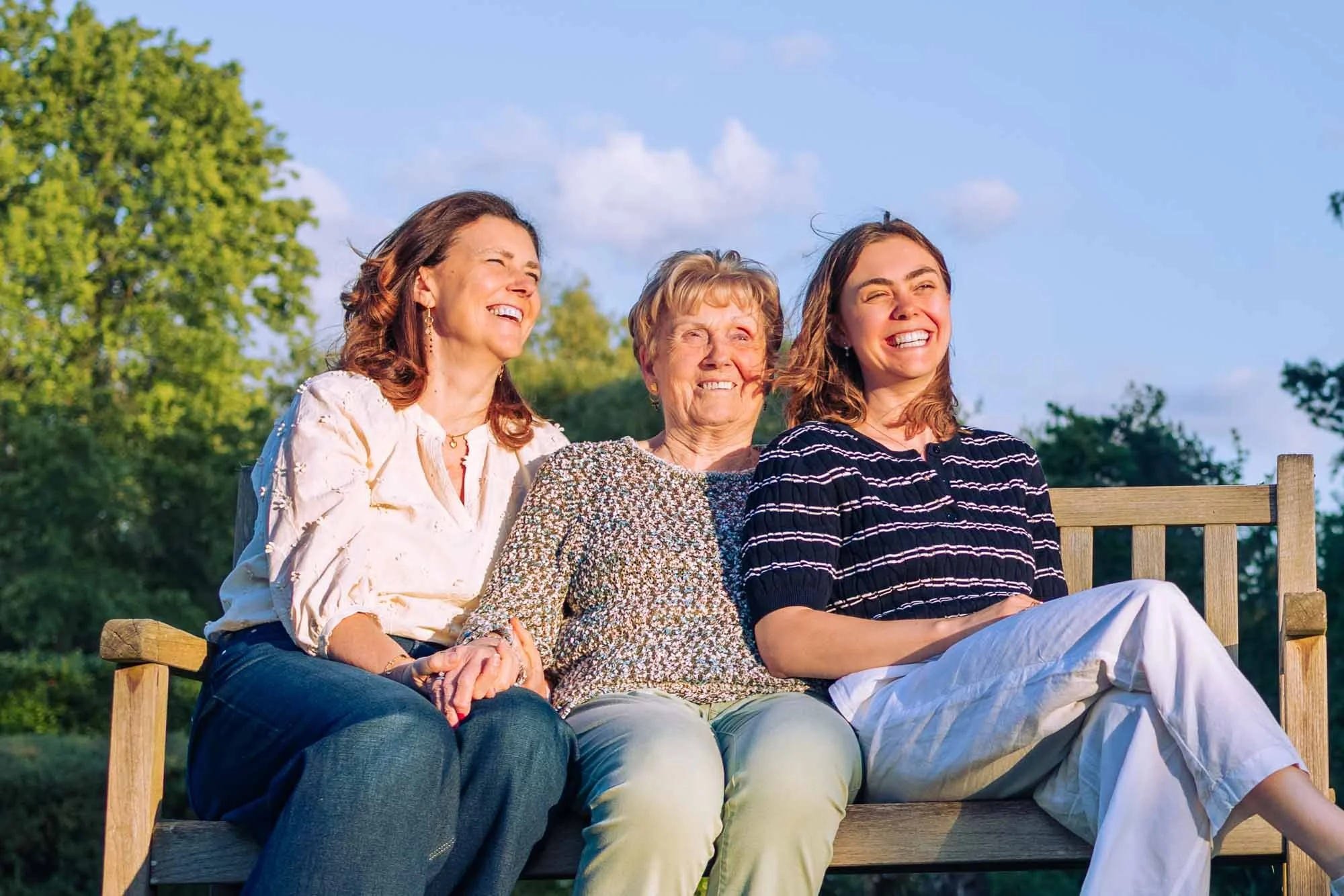 "Amber Sels met haar mama en grootmoeder op een houten bank, samen lachend in het avondlicht in een groene tuin in Rijmenam"
