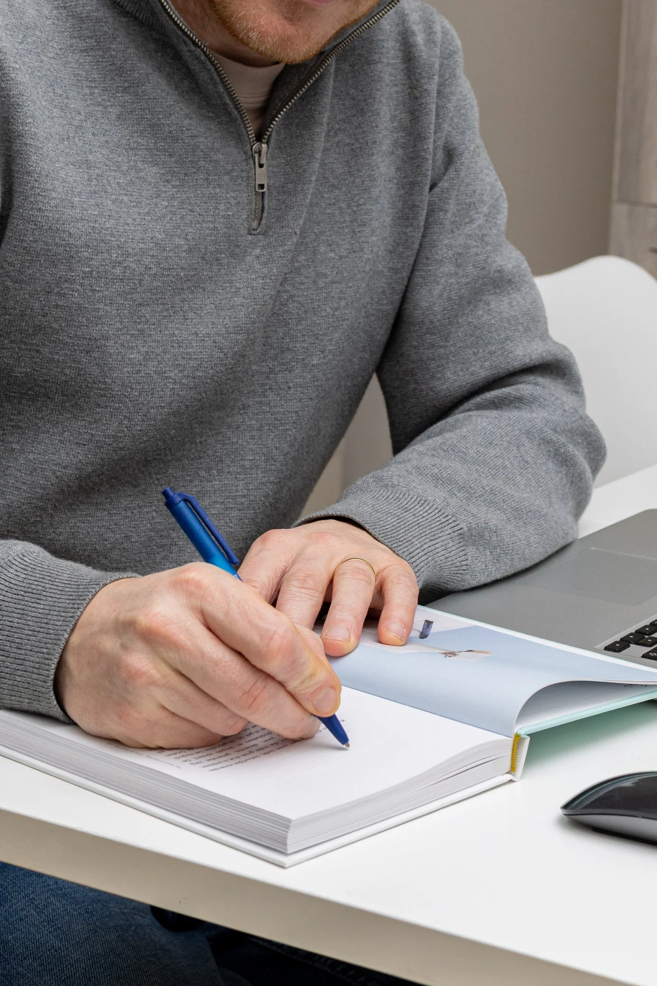A person wearing a gray zip-up sweater sitting at a white desk, writing in a book with a blue pen. There is a laptop and a computer mouse on the desk.