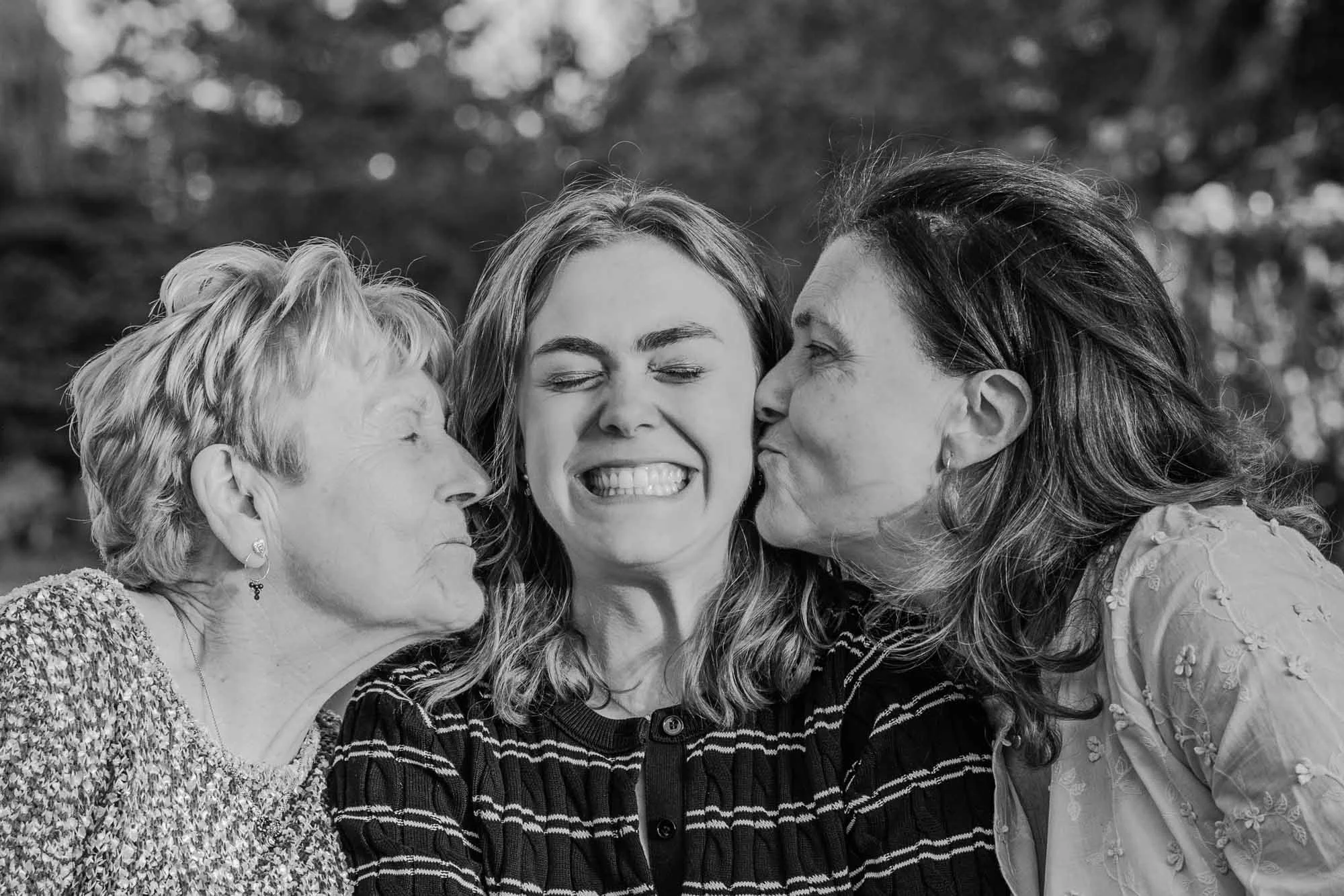 "Zwart-wit foto van Amber Sels tussen haar mama en grootmoeder, die haar elk een kus op de wang geven – spontaan familieportret met drie generaties"