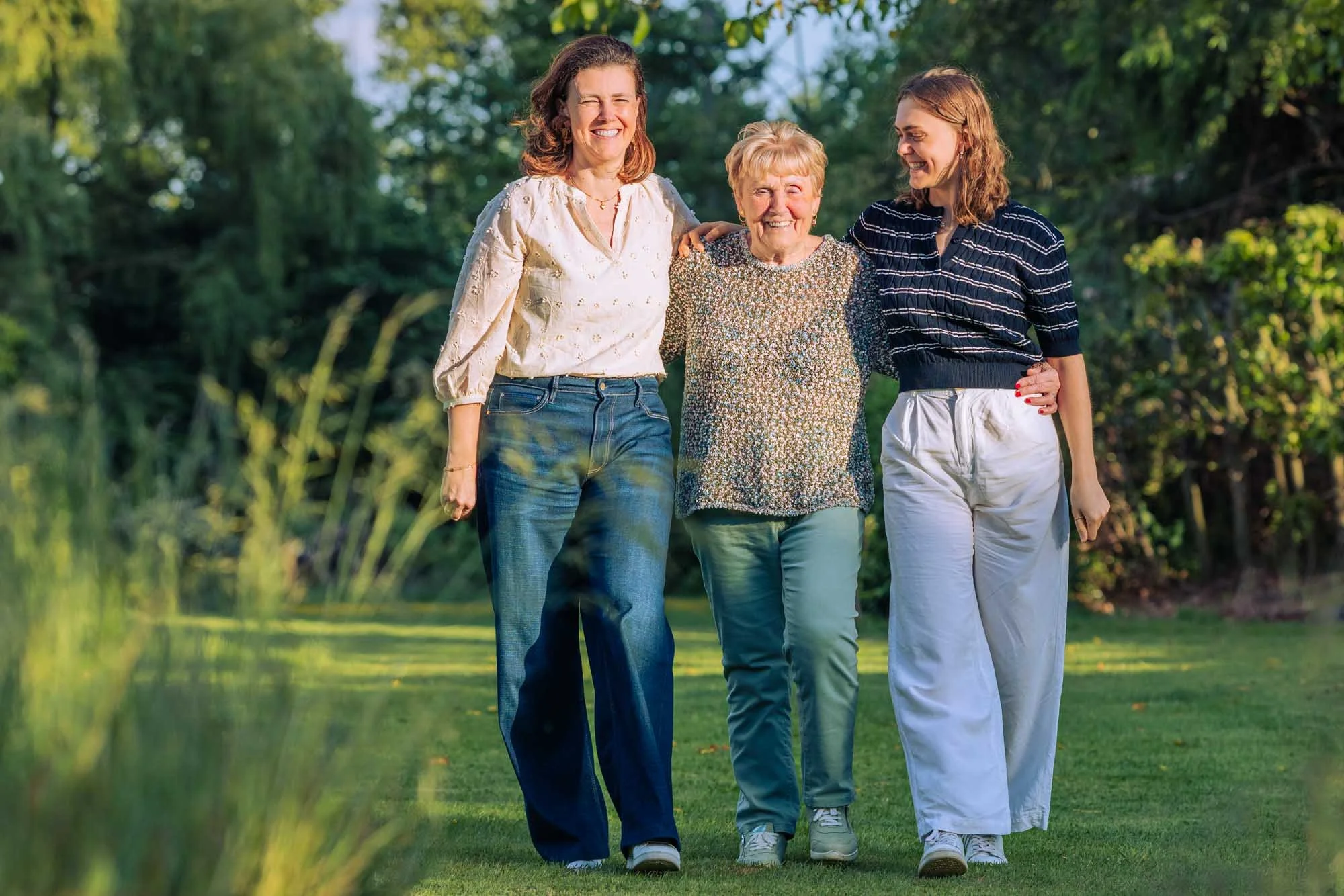 "Drie generaties vrouwen – Amber Sels, haar mama en grootmoeder – wandelend en lachend in een groene tuin in Rijmenam, lifestyle familieportret"