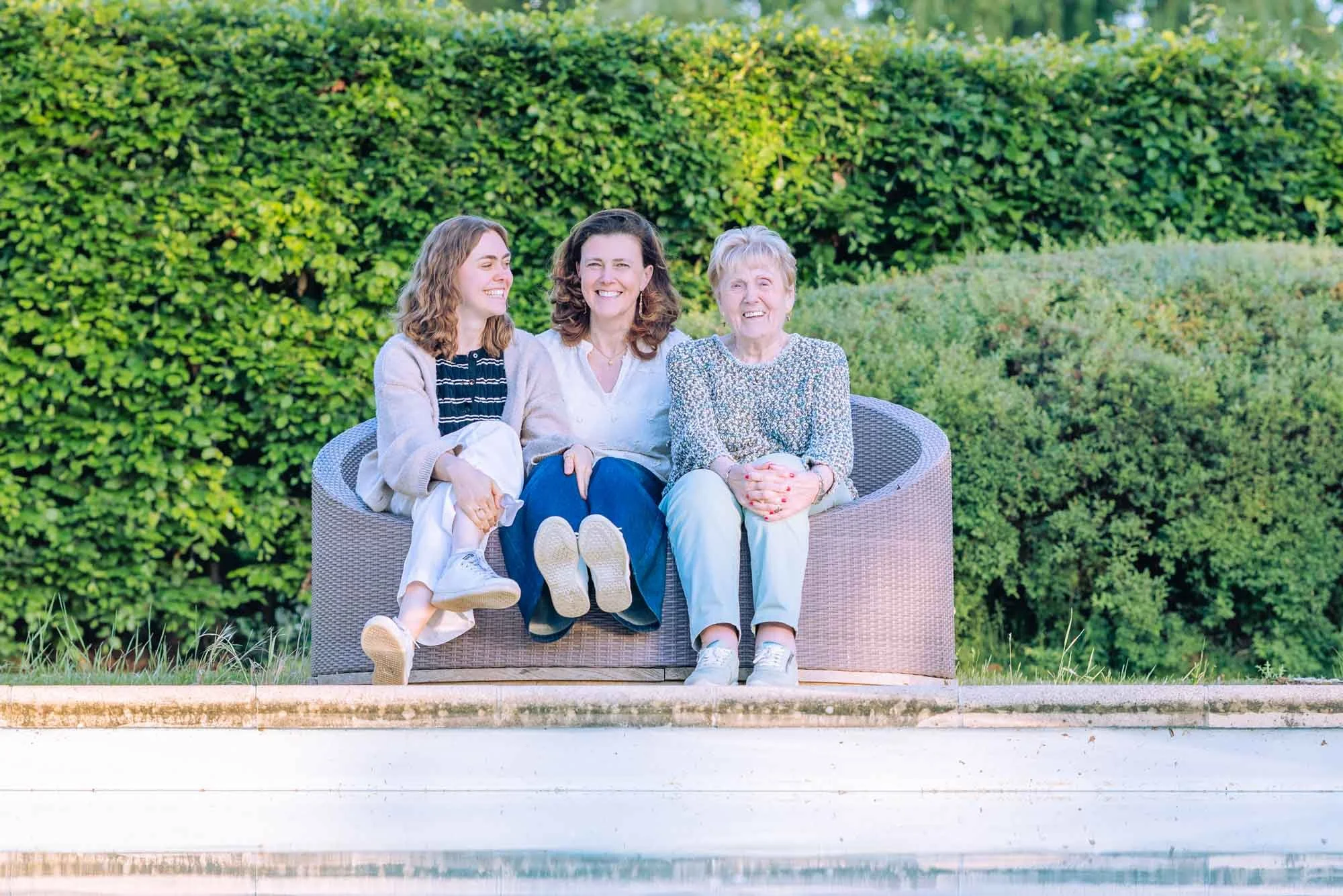 "Amber Sels, haar mama en grootmoeder zittend op een ronde buitenzetel voor een haag, ontspannen familiemoment tijdens een lifestyle portretsessie"