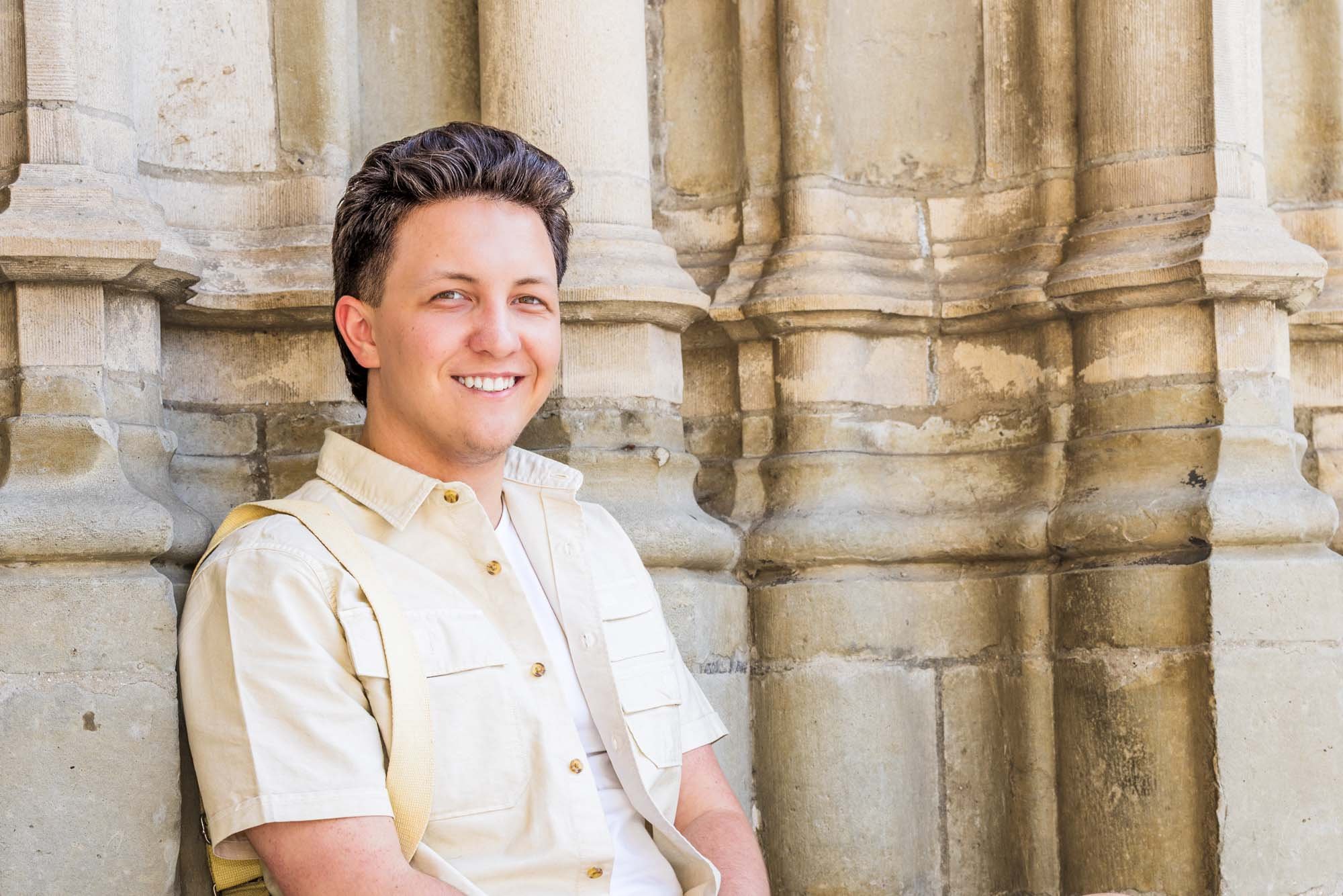 Lachend portret van Alex Verspecht zittend tegen een zandkleurige gevel van een kerk in Mechelen, zomerse sfeer. Locatie: sint Rombouts toren 
