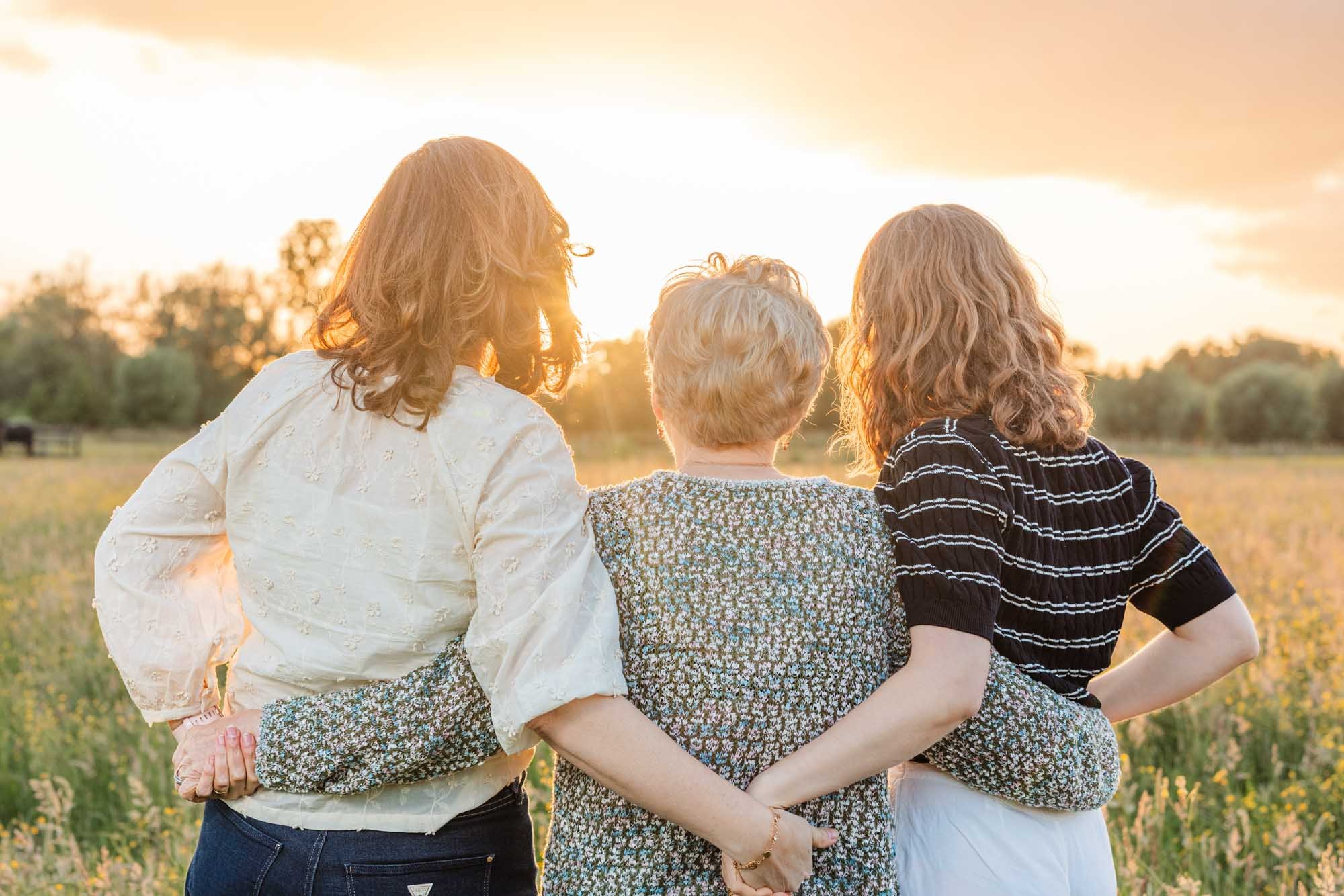 Drie generaties vrouwen delen een warme omhelzing in een bloemenveld bij zonsondergang. Gezinsportret door Leysen Photography met natuurlijke sfeer en gouden licht.