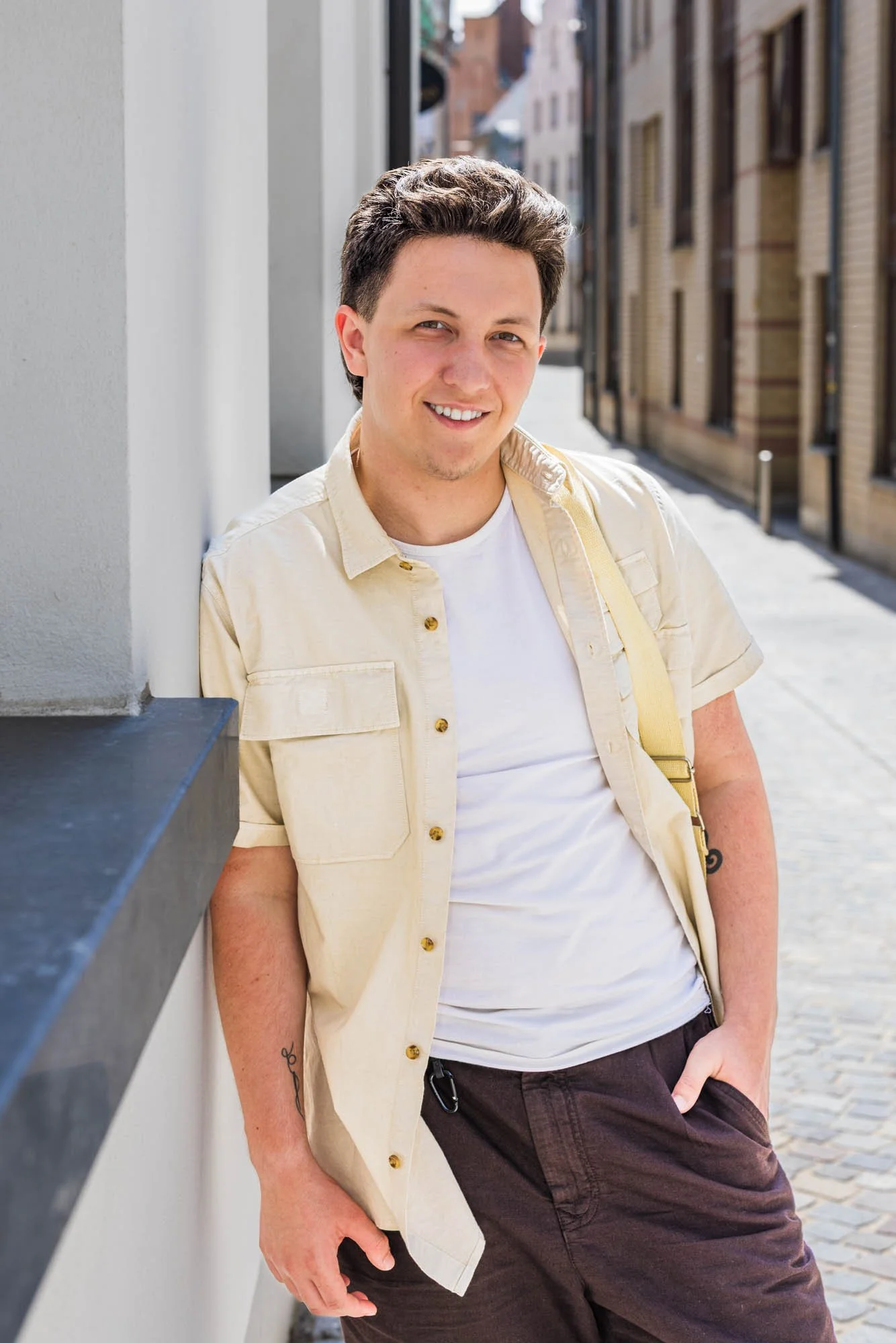 Casual portret van Alex Verspecht leunend tegen een witte muur in een smal straatje in Mechelen, met zonlicht op zijn gezicht"
