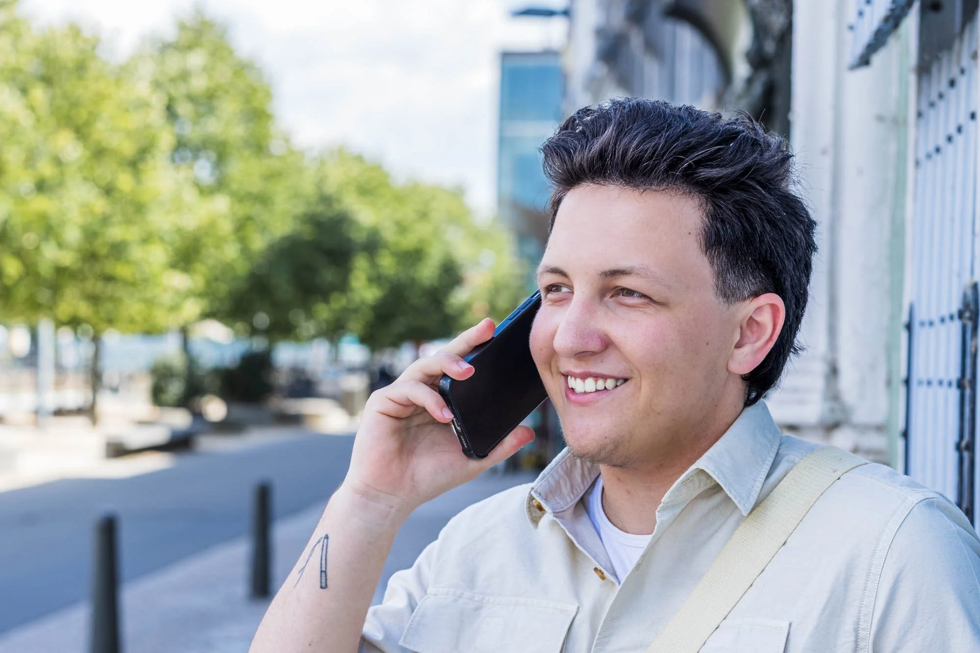 Portretfoto van Alex Verspecht al bellend op straat in Mechelen, met zomerse achtergrond van bomen en moderne gebouwen"