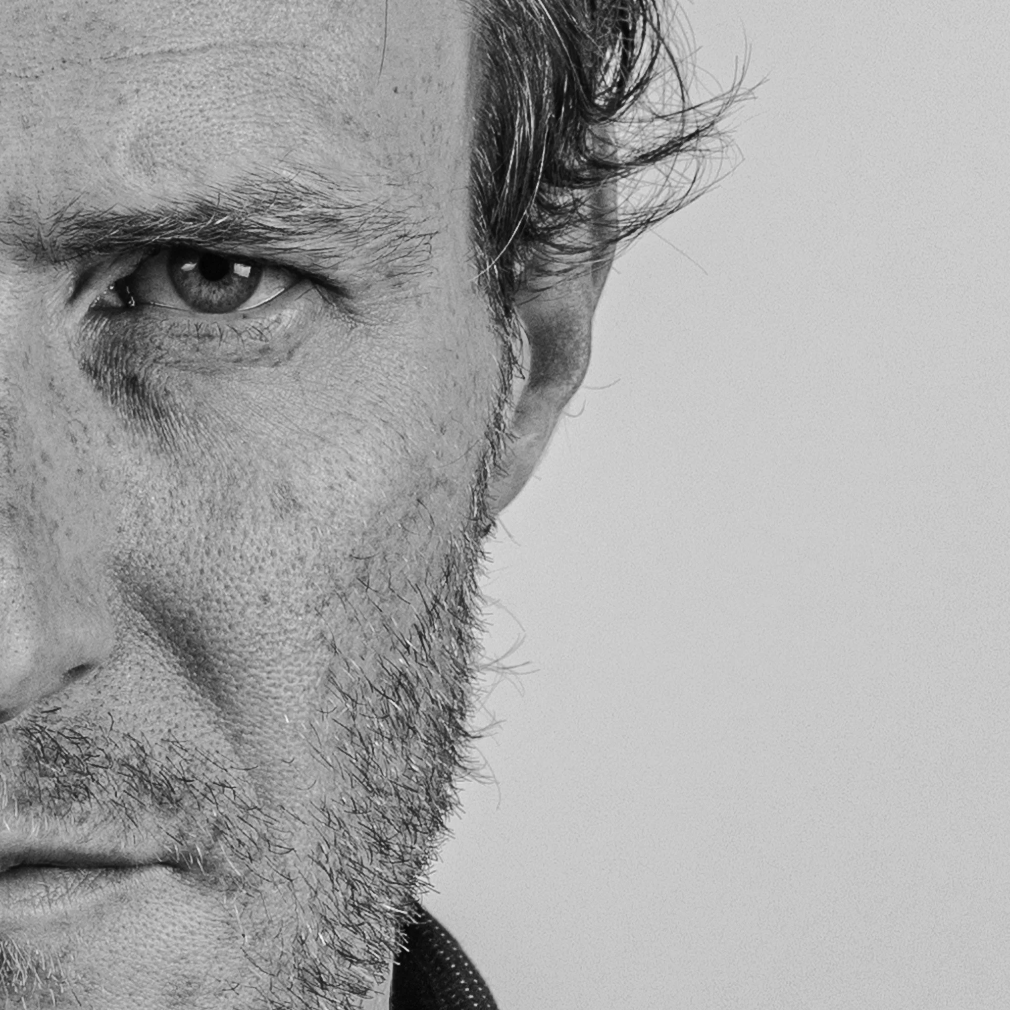 Close-up black and white photograph of a man's face, focusing on his left eye and part of his cheek, with a textured background.