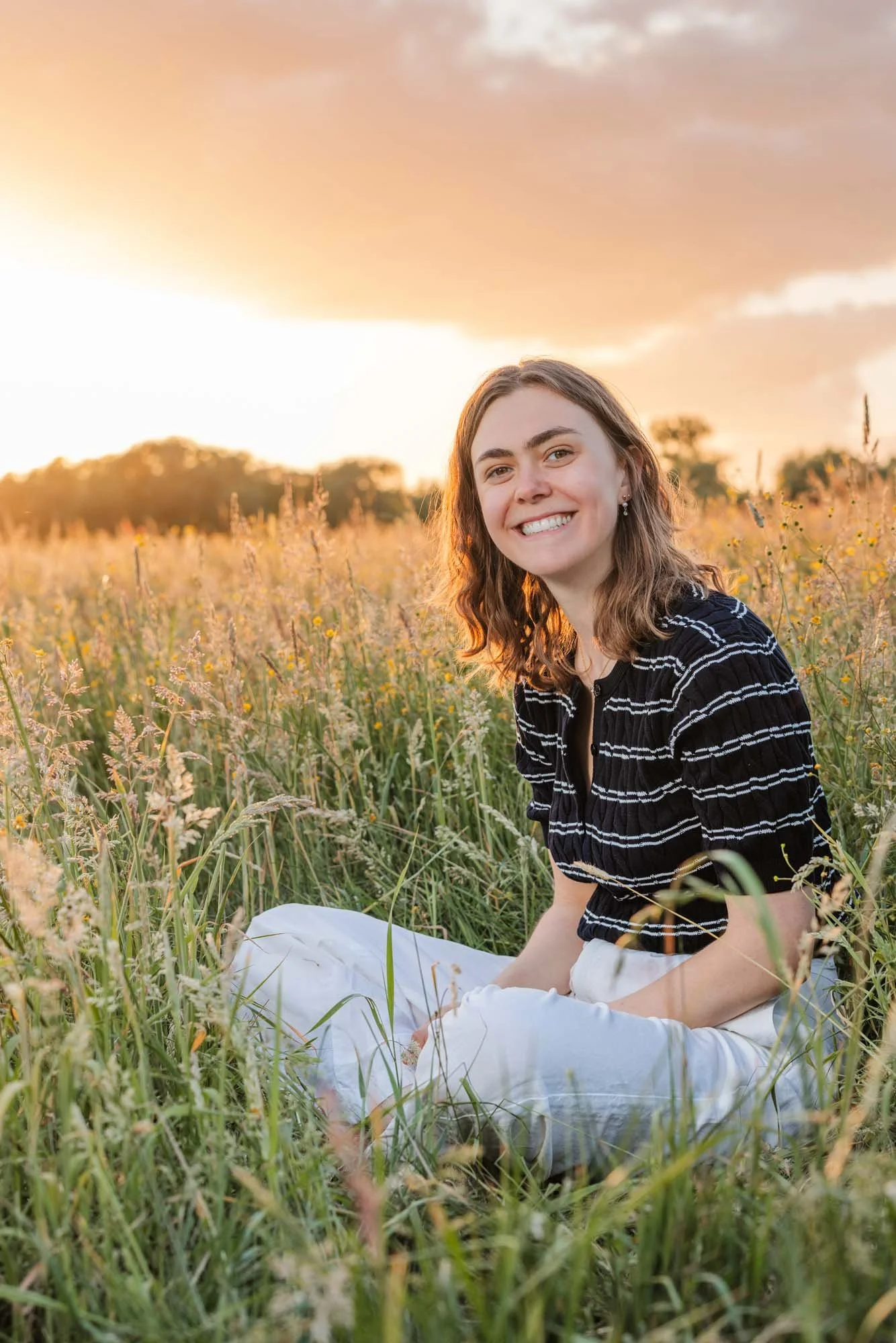 "Amber Sels zit glimlachend in een bloemenveld bij zonsondergang, omringd door lang gras en een warme gouden gloed – zomers lifestyle portret"
