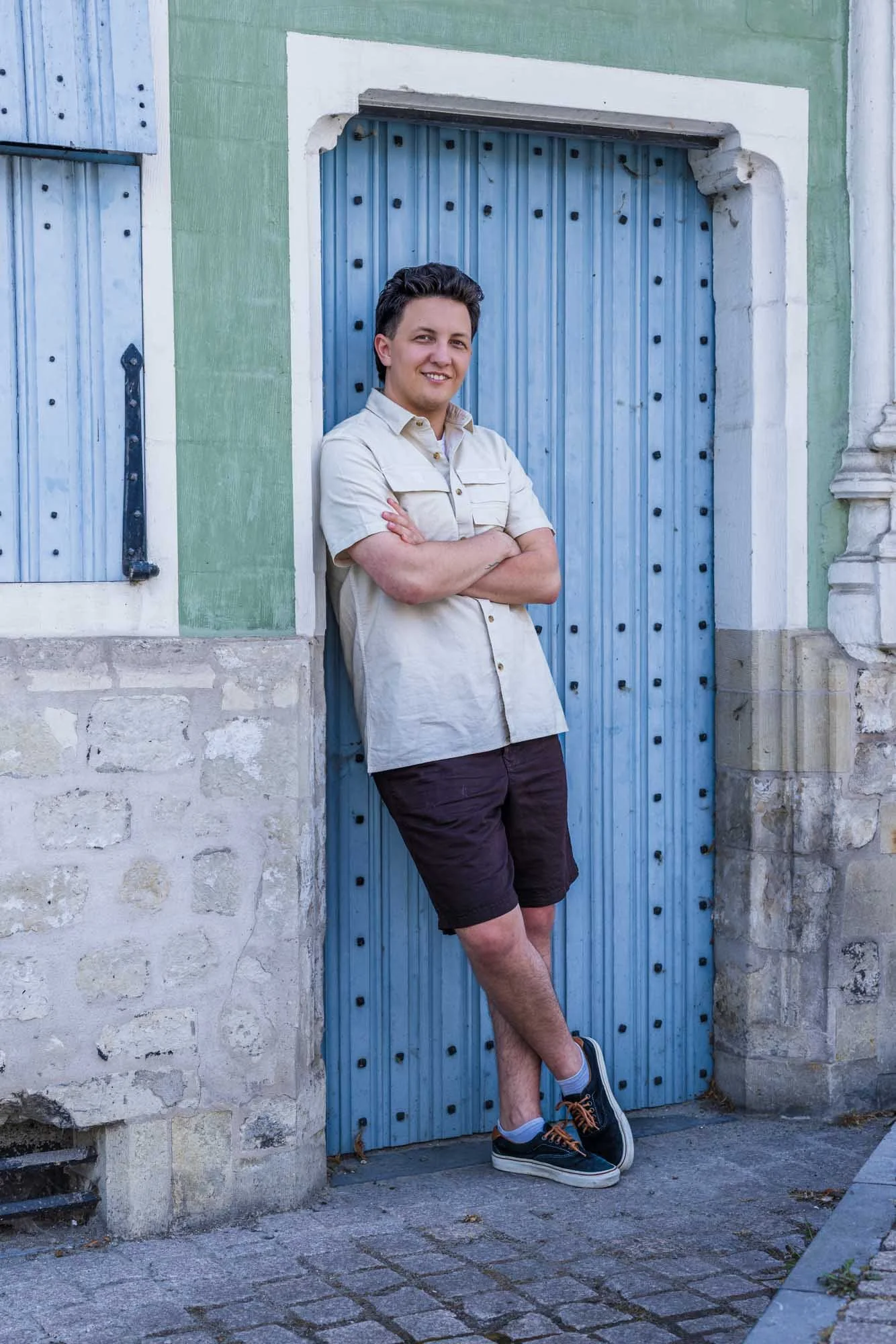 alt="Volledig portret van Alex Verspecht poserend met gekruiste armen voor een turquoise gevel en blauwe deur in Mechelen, casual stijl"
