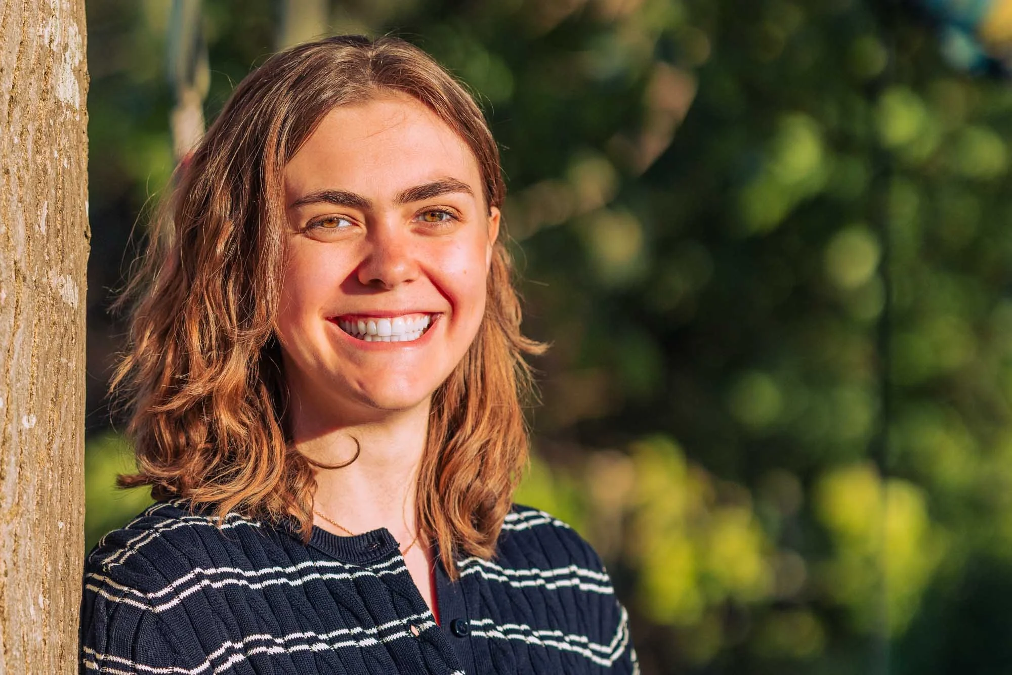 "Portret van Amber Sels die breed glimlacht terwijl ze leunt tegen een boomstam, genomen in natuurlijk avondlicht in Rijmenam"
