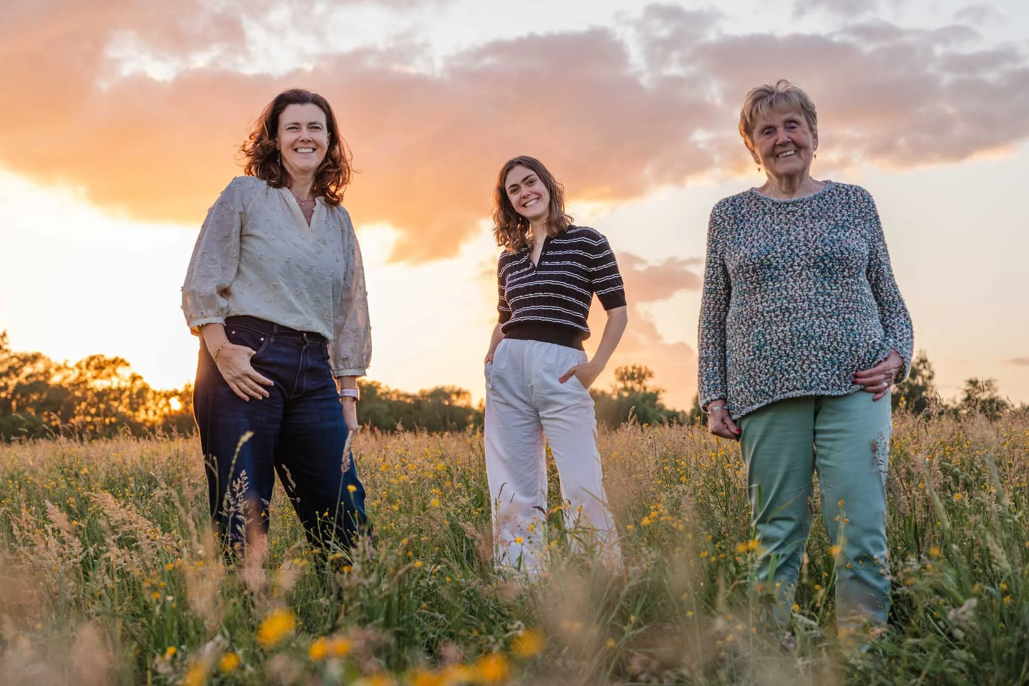 Moeder, dochter en grootmoeder staan zij aan zij in een bloemenveld bij zonsondergang. Portretfotografie door Leysen Photography met natuurlijke expressie en warme kleuren.
