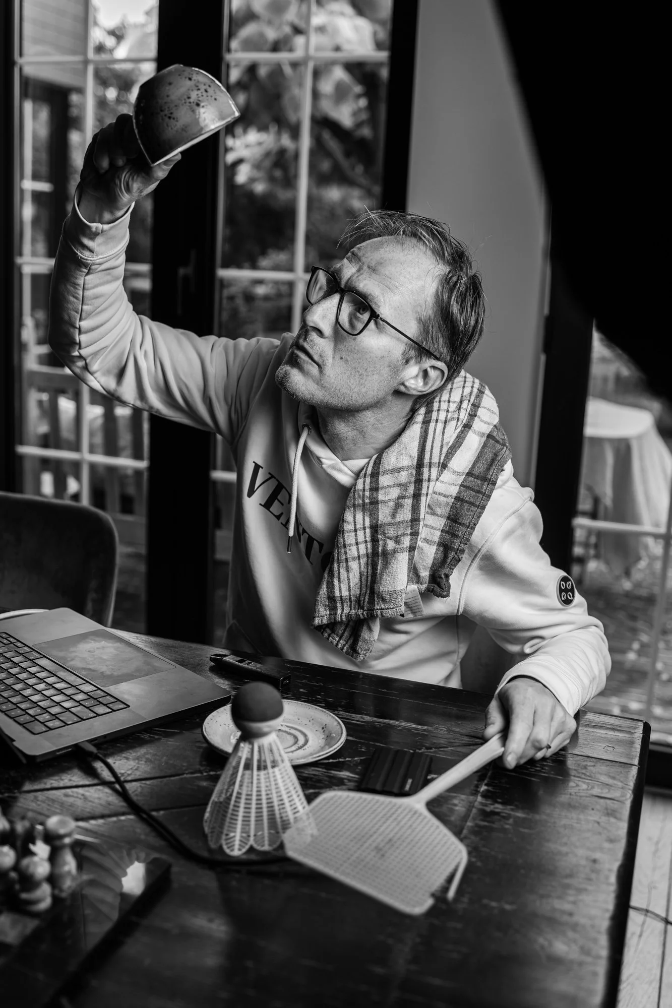 A man with glasses and a plaid towel over his shoulder sitting at a dining table, holding a slice of watermelon in the air, with a laptop, badminton racket, and shuttlecock on the table, near a window.